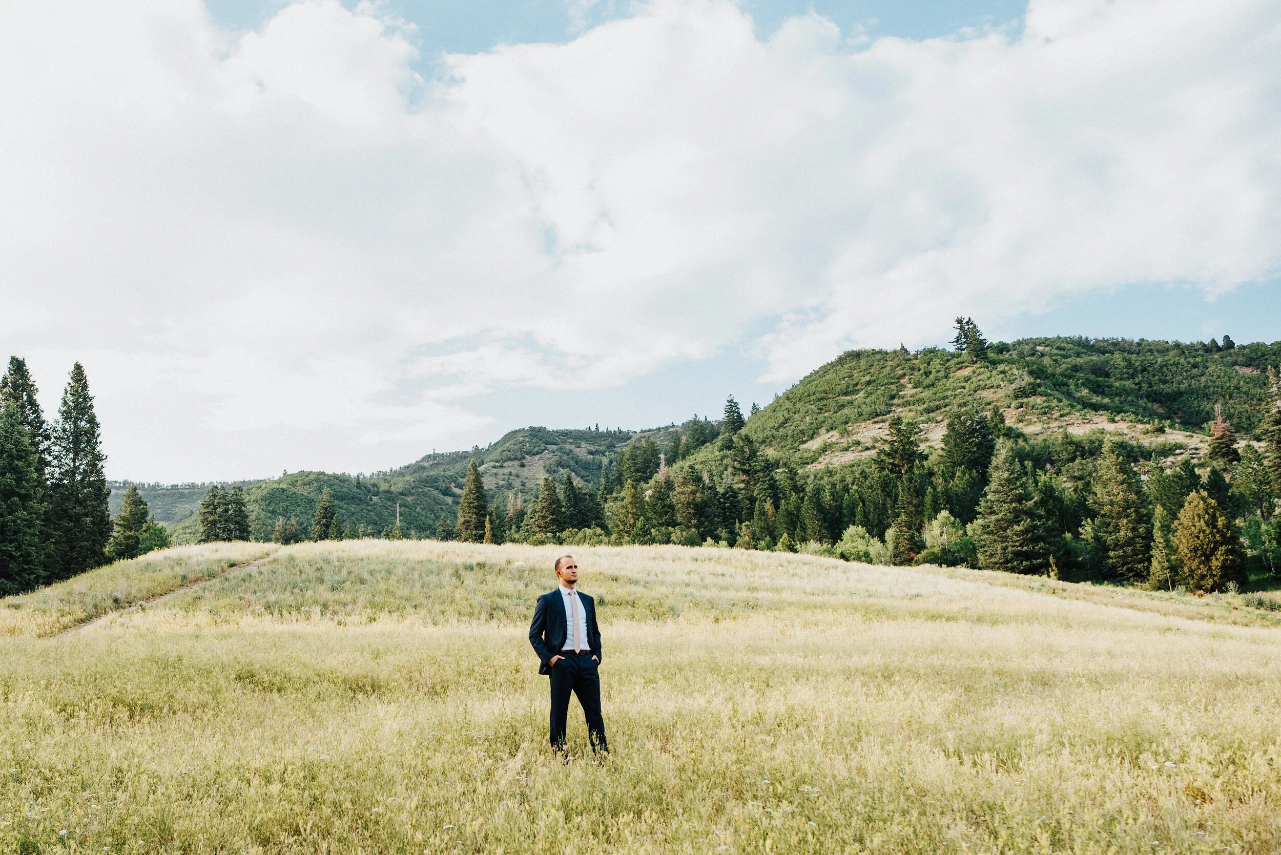 A handsome groom strikes a confident pose looking off into the distance with hands in his pockets in the middle of a grassy field. American Fork Canyon groom individual photo powerful pose navy suit light pink tie tall grass small mountains evergree…