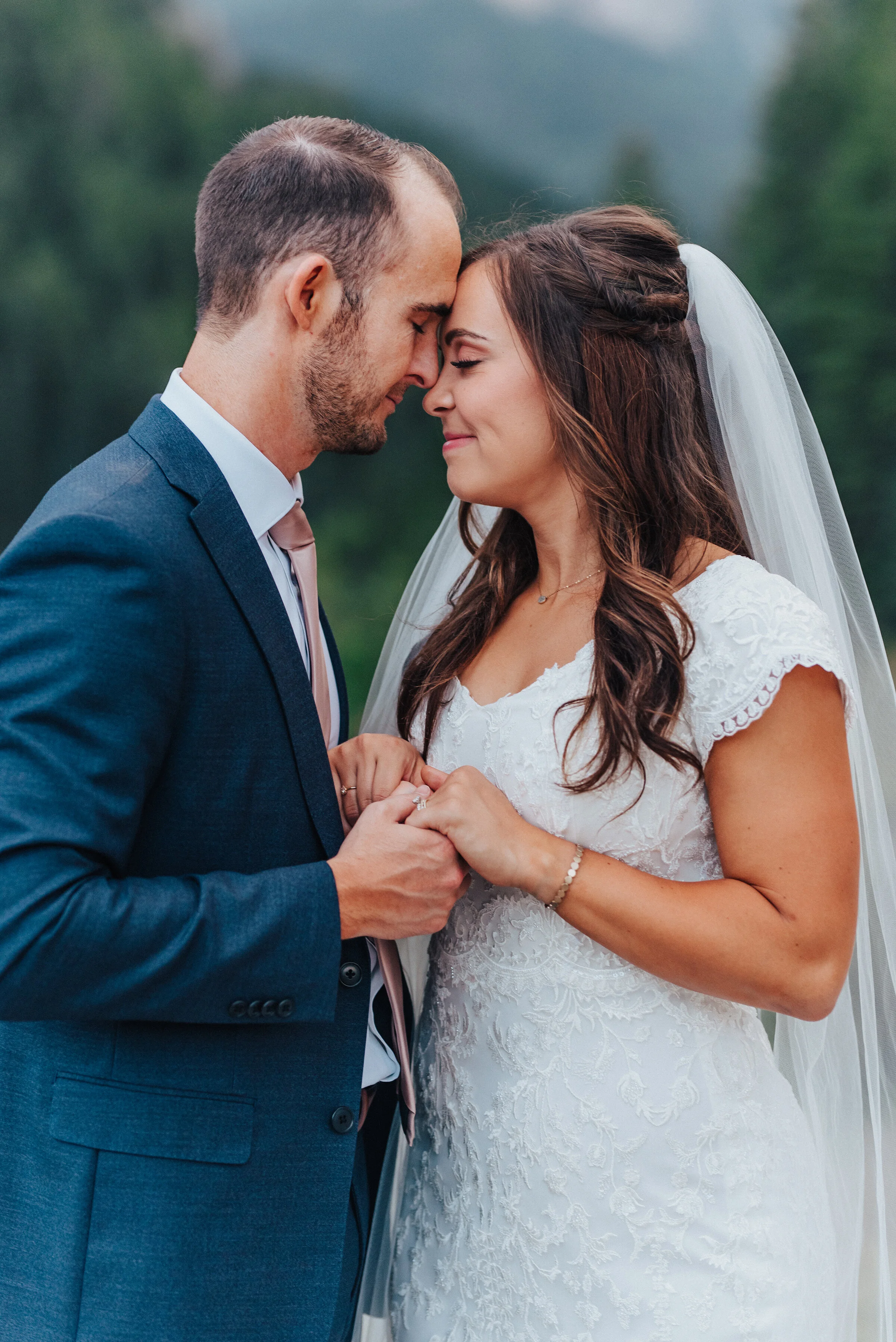 A bride and groom softly smile while resting their foreheads on each other and holding hands at Tibble Fork Reservoir. Holding hands navy suit light pink tie long veil closed eyes floral design dress evergreen trees in the background curly hair #hus