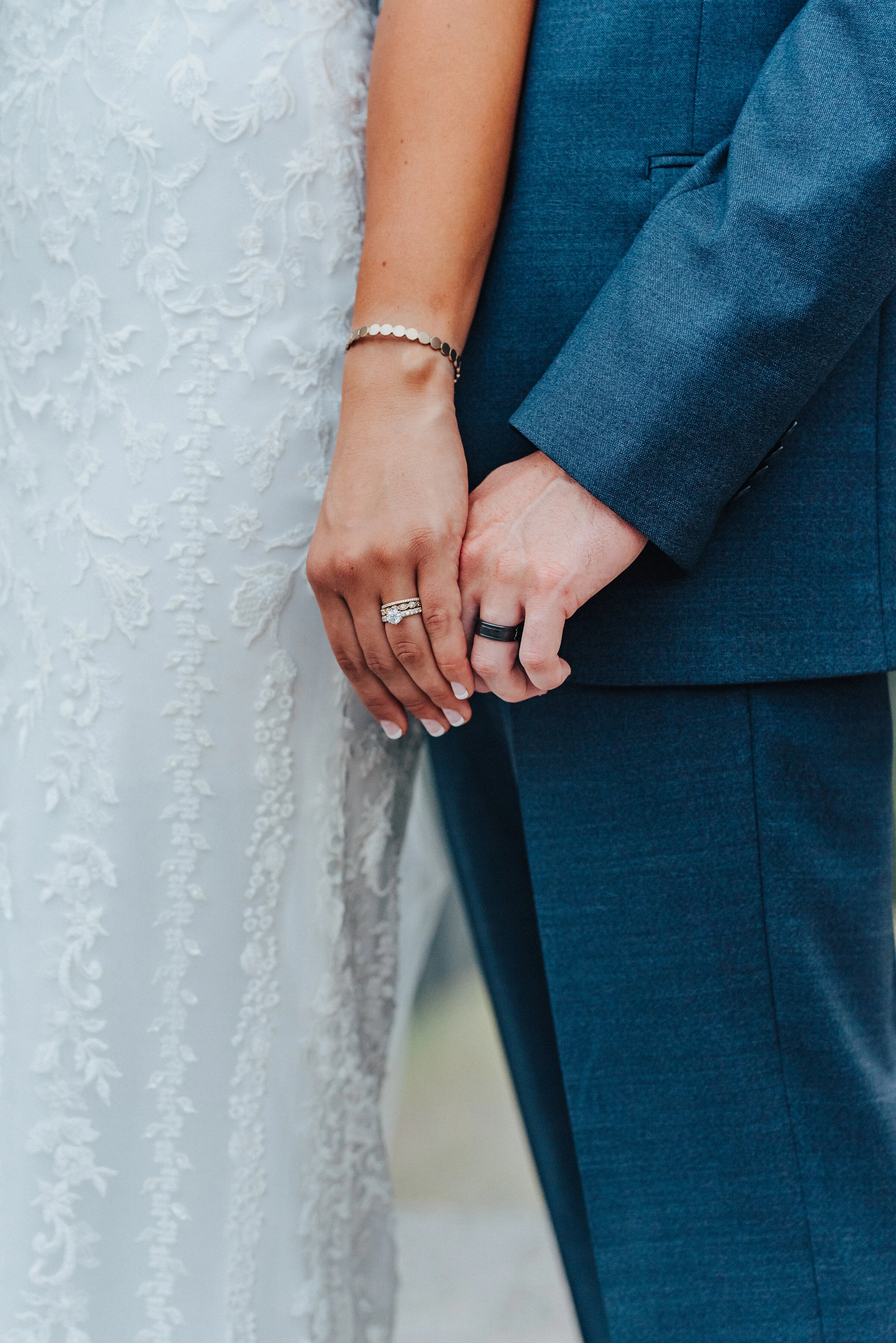  A close-up of the bride and groom’s hands touching while showing off their gorgeous rings. Almost interlocked hands navy blue suit french tip nails close-up shot in American Fork floral dress design dark ring for the groom bride in front of groom #h