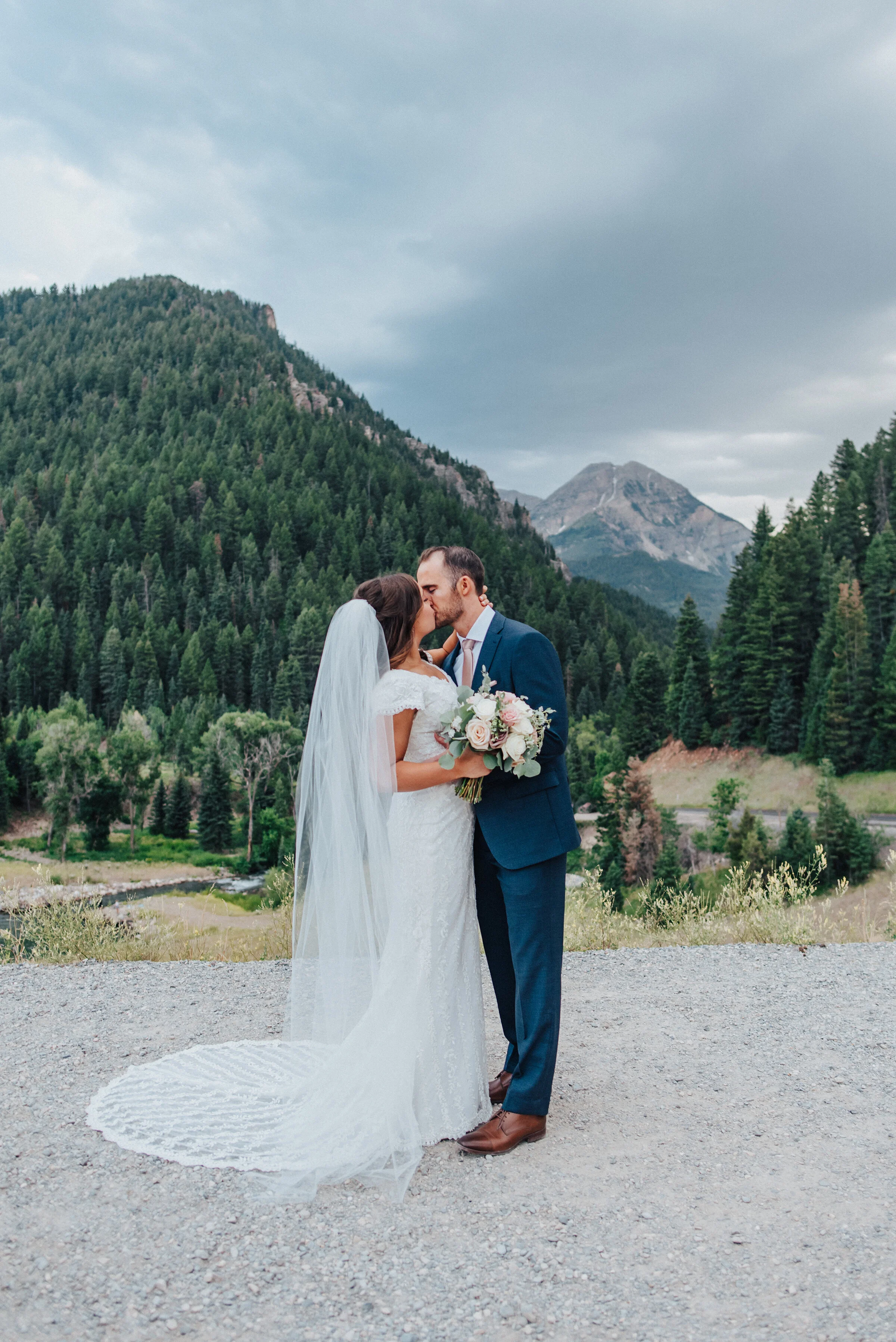  A beautiful couple shares a kiss while standing on a smooth sidewalk that surrounds the breathtaking Tibble Fork Reservoir. Navy suit light pink tie light pink floral bouquet exchanging glances evergreen trees mountains in the background #husbandand