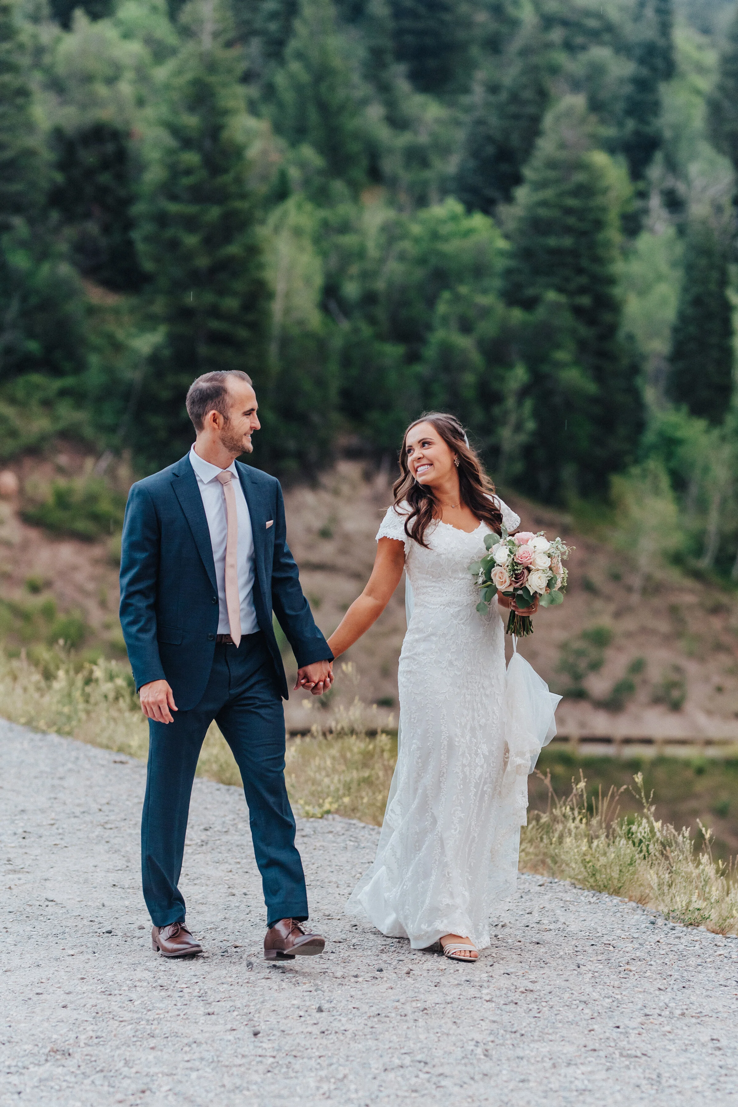  A smiling couple stroll down a paved pathway that surrounds the Tibble Fork Reservoir in American Fork canyon while holding hands. Navy suit light pink tie light pink floral bouquet exchanging glances evergreen trees #husbandandwife #younglove #tibb