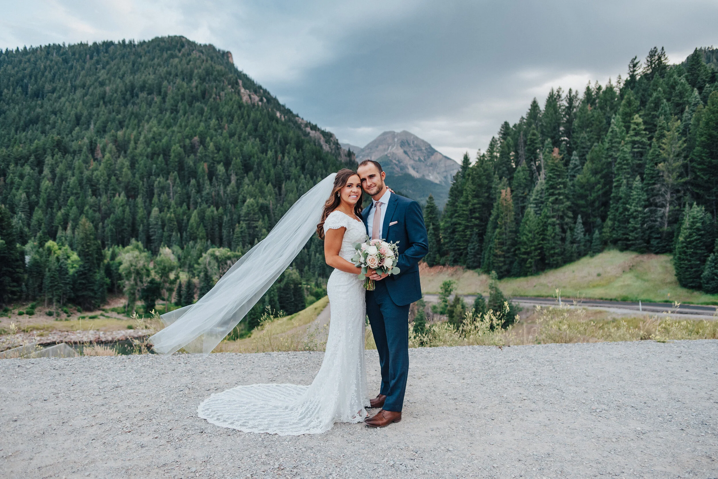 A gorgeous bride and groom look into the camera while softly embracing each other in front of the breathtaking Tibble Fork Reservoir. Navy suit light pink tie light pink floral bouquet evergreen trees mountains in the background classic wedding shot…