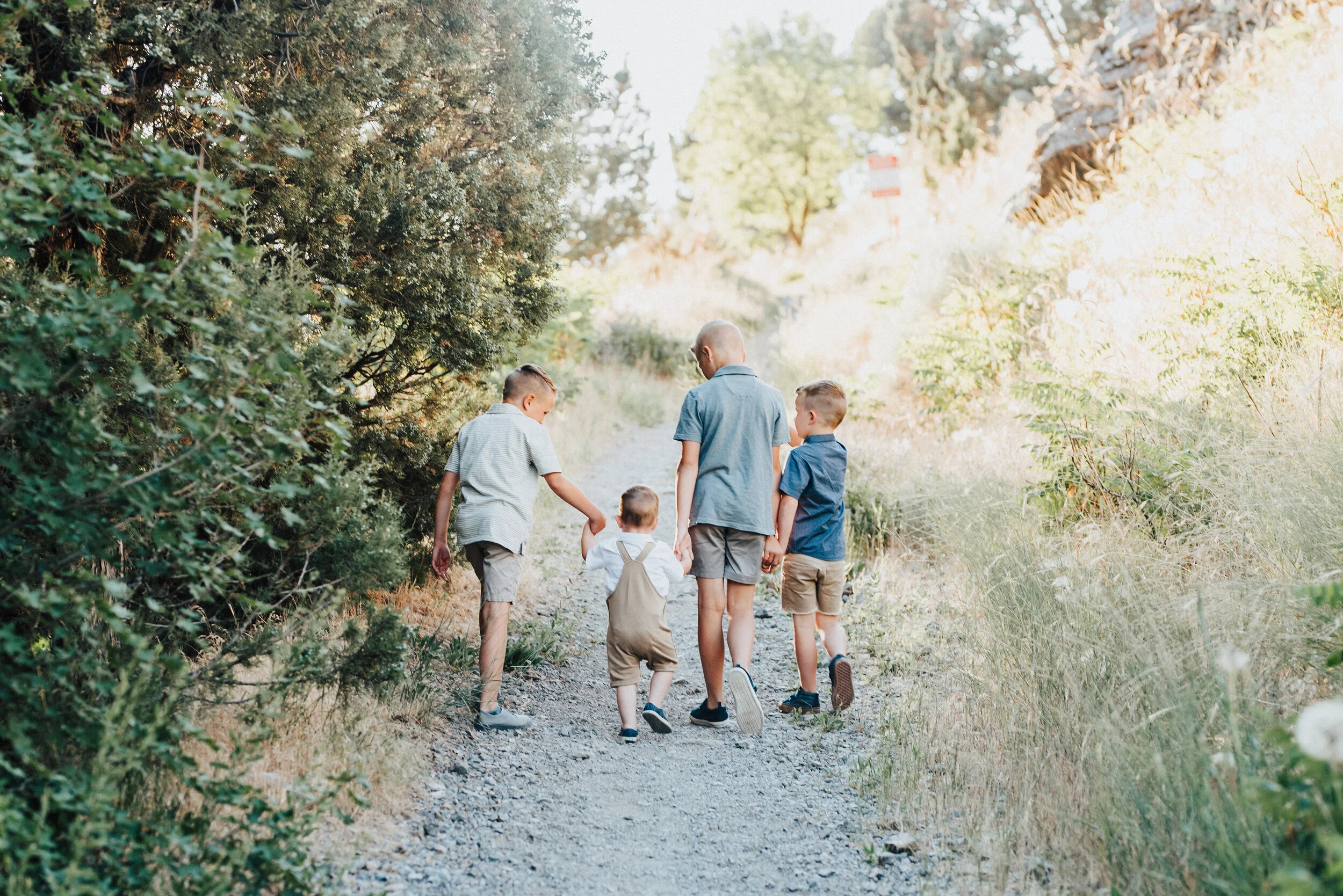 Photo captured from behind of four young brothers holding hands strolling down a sunlit trodden path in Providence Canyon. Color-coordinated light blue and khakis brotherly love walking down a path outdoor family photos #providencecanyon #utahphotog…