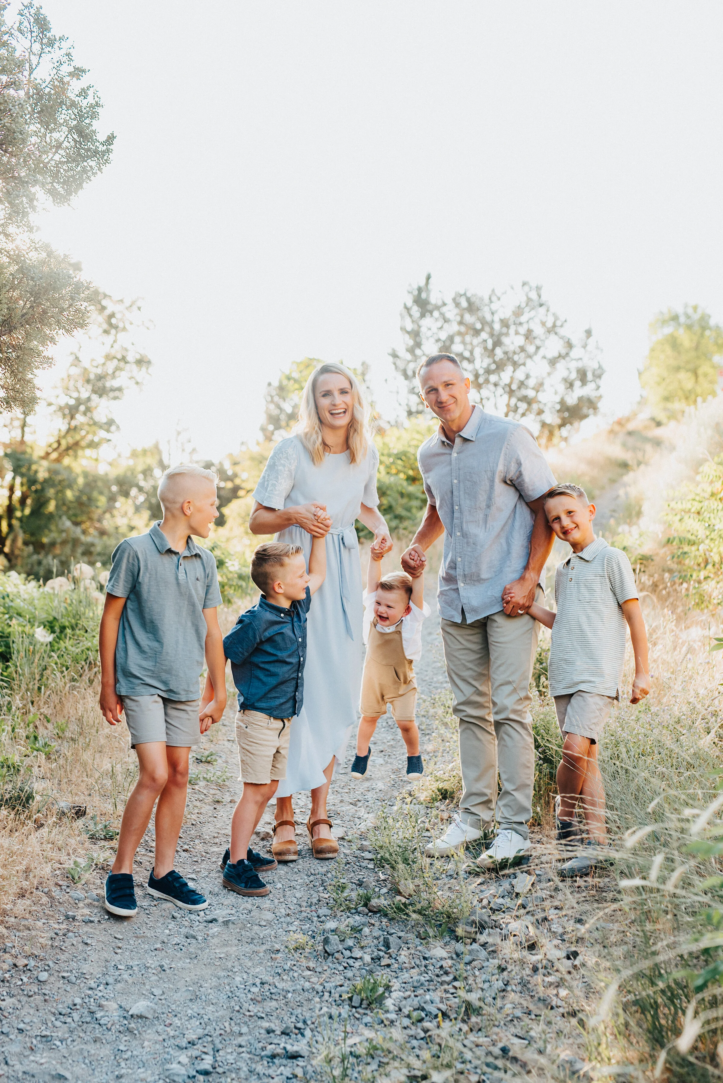  An adorable smiling family of six are holding hands while walking down a trodden footpath in Providence Canyon, UT. Light blue and khakis soft outdoor setting family photoshoot backlit sunlight in utah dessert #providencecanyon #utahphotography #uta