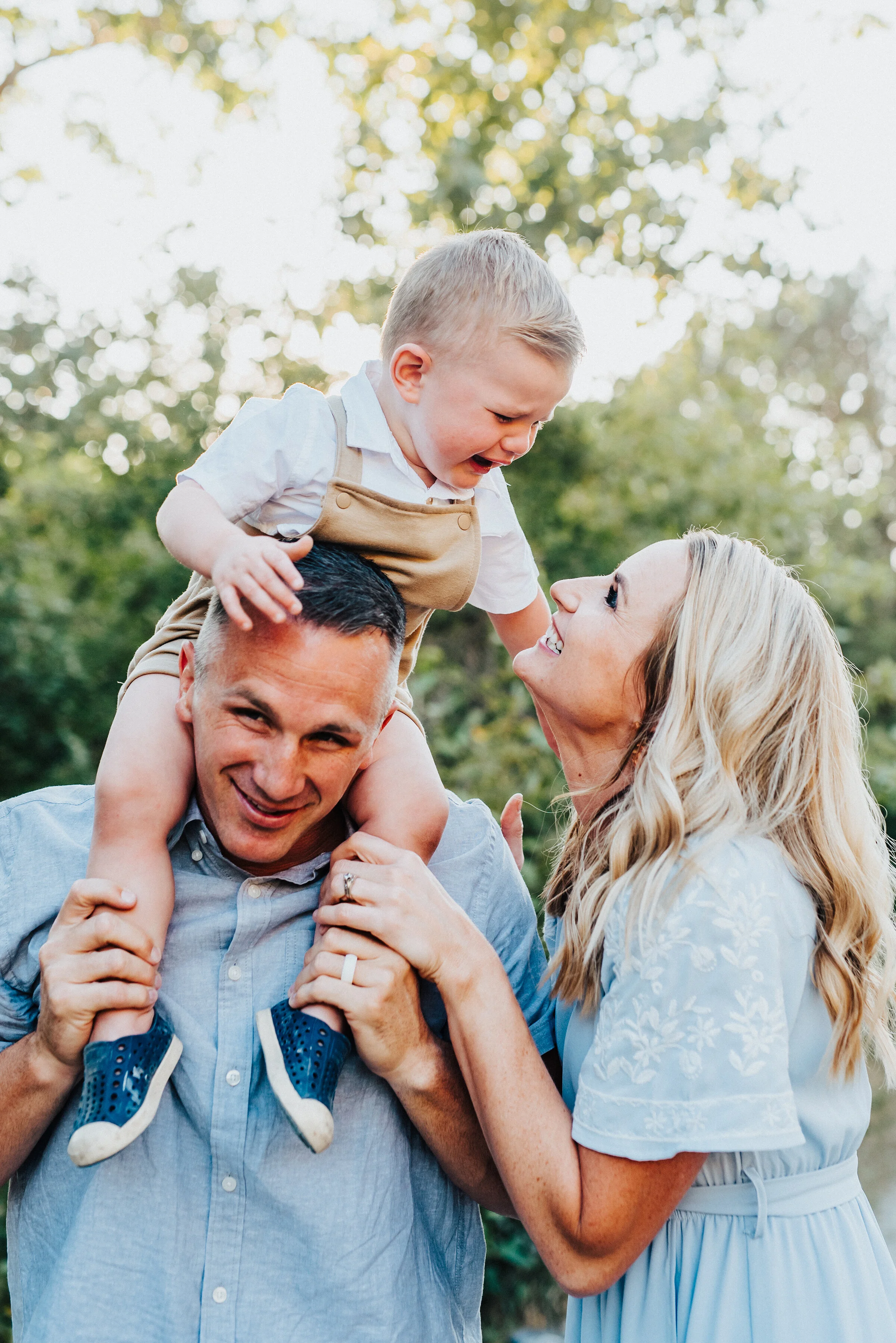  Two smitten parents are playfully posing with their youngest son, who finds the experience a little less than enjoyable on his dad’s shoulders without any loss of adorableness. Light blue and khakis soft outdoor setting family photoshoot Providence 