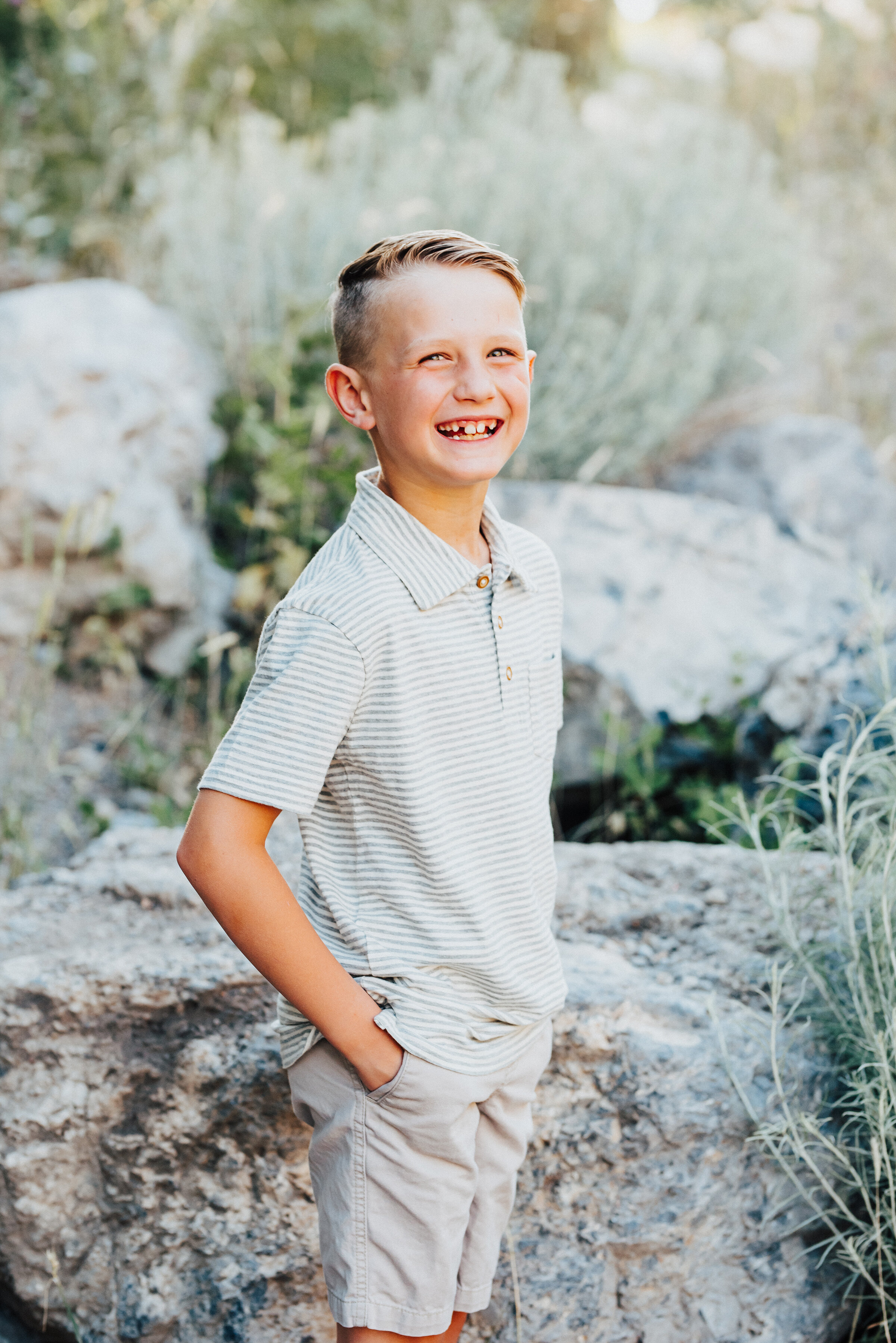  An adorable young boy cheesily grins into the camera with hands in his pockets while standing near a boulder in Providence Canyon, UT. light blue and khakis soft outdoor setting casual standing cheesy toothy grin #providencecanyon #utahphotography #