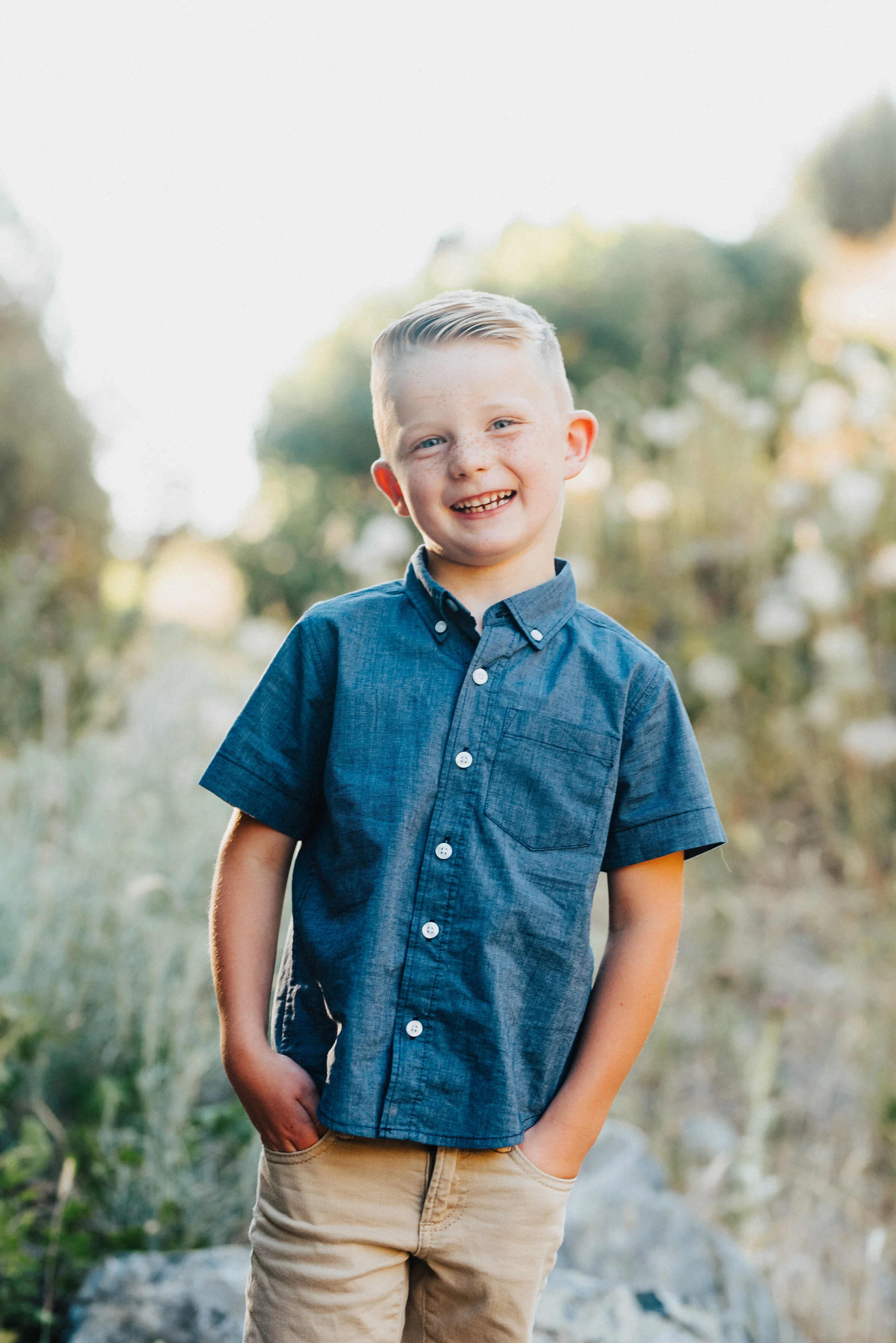  A big smile is spread across a young boy’s face while his hands rest in his pockets in the canyon. medium blue and khakis soft outdoor setting tilted head hands in his pockets sunlit outdoor backdrop #providencecanyon #utahphotography #utah #familyp