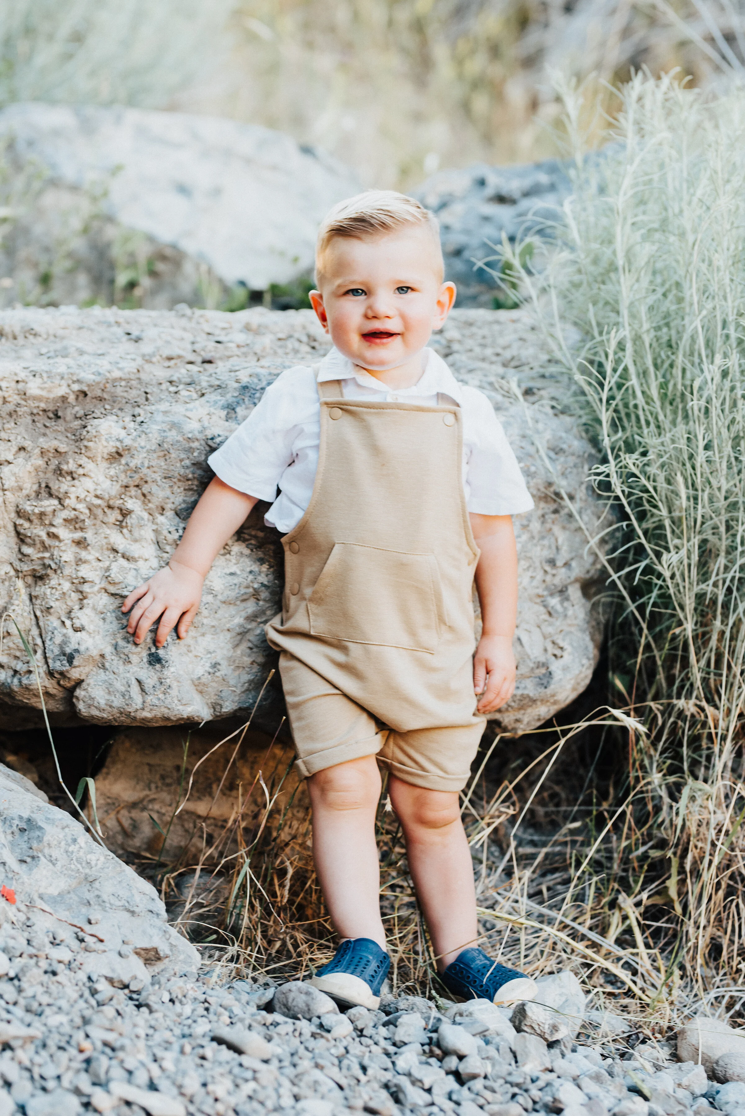  An adorable toddler in precious tan overalls with perfectly combed hair rests up against a large boulder in Providence Canyon, UT. toddler wearing overalls light blue and khakis soft outdoor setting standing on rocky path #familylove #photoshoot #fa