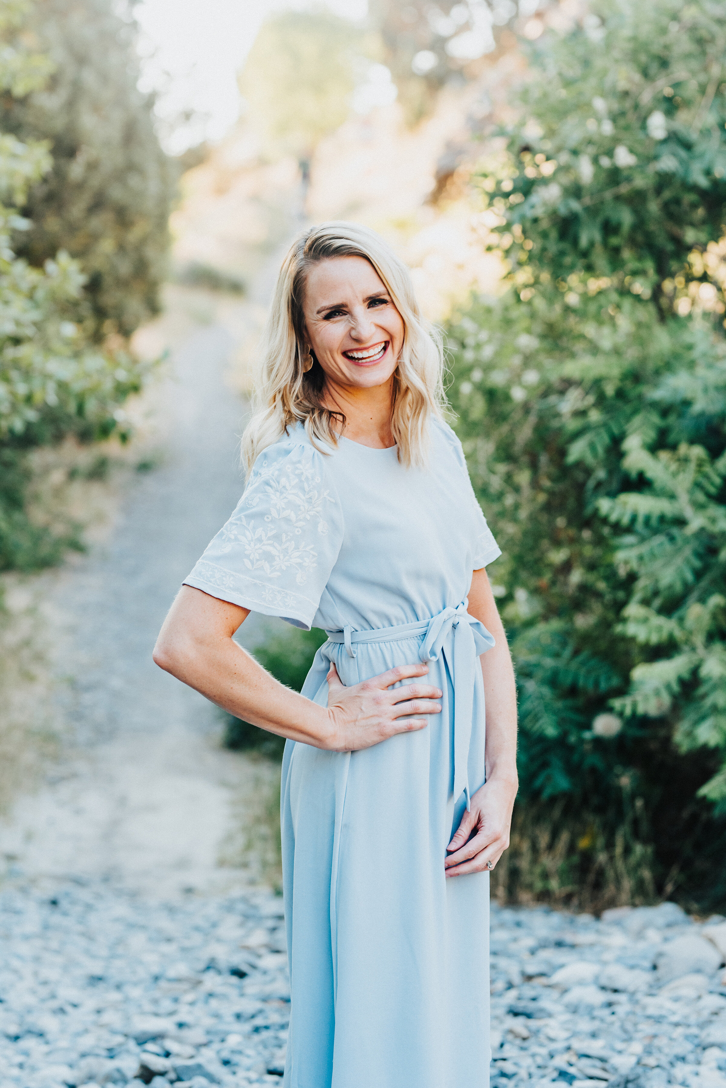  Beautiful mom has her hand hugging her hip as she gives a big laughing smile to the camera. Light blue dress with fluttery sleeves standing on the stony pathway in providence canyon utah #providencecanyon #utahphotography #utah #familyphoto #family 