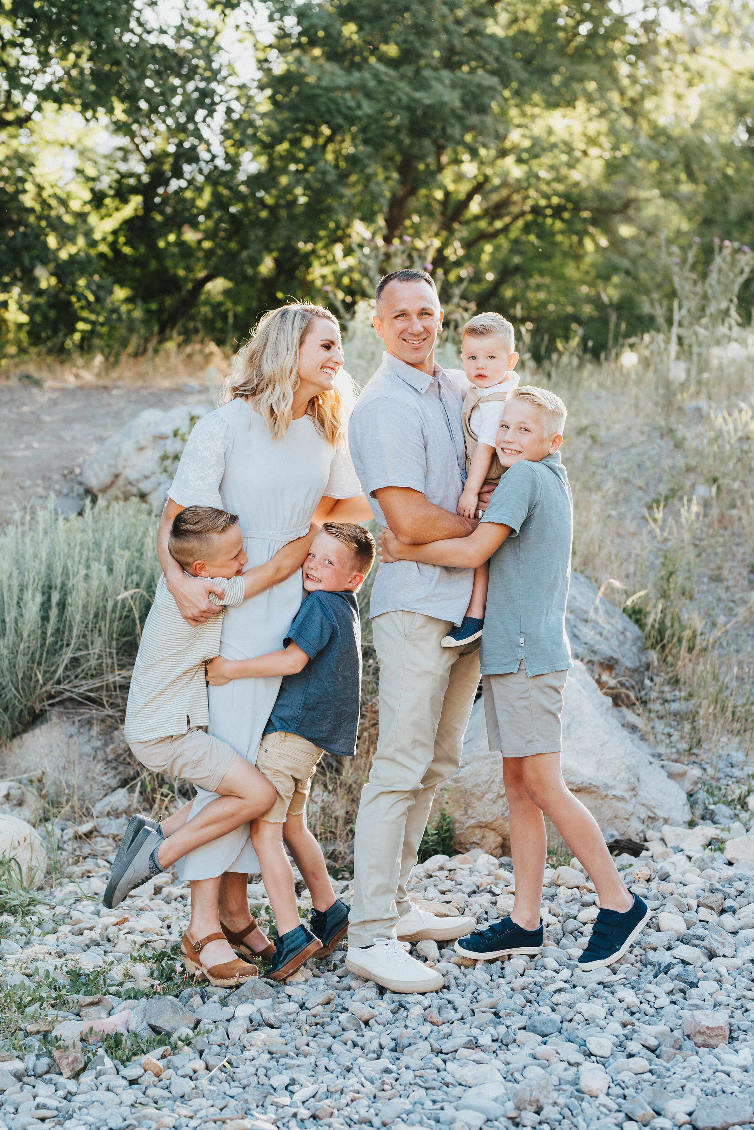  Beautiful family is giving each other tight hugs with the sun peeking out from behind the trees. Light blue and khakis soft outdoor setting looking at different directions family all together #providencecanyon #utahphotography #utah #familyphoto #family #familyphotography #familyphotographer #outdoorphotoshoot #hugs #familyhugs #familyhugging #sillyhugs 