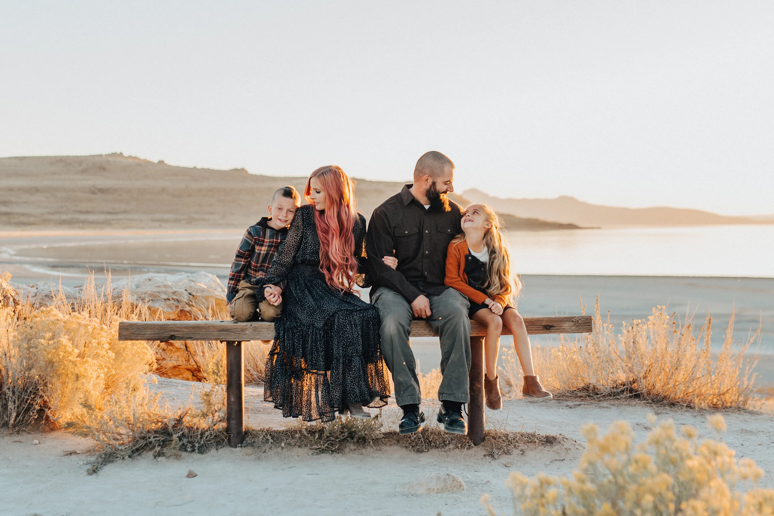  A glowing family sitting together in a beautiful sun set photo shoot by Kristi Alyse Photography. Utah photographer goals ideas and inspiration Antelope Island photo shoot goals young family photo shoot inspiration sitting family pose inspiration lo