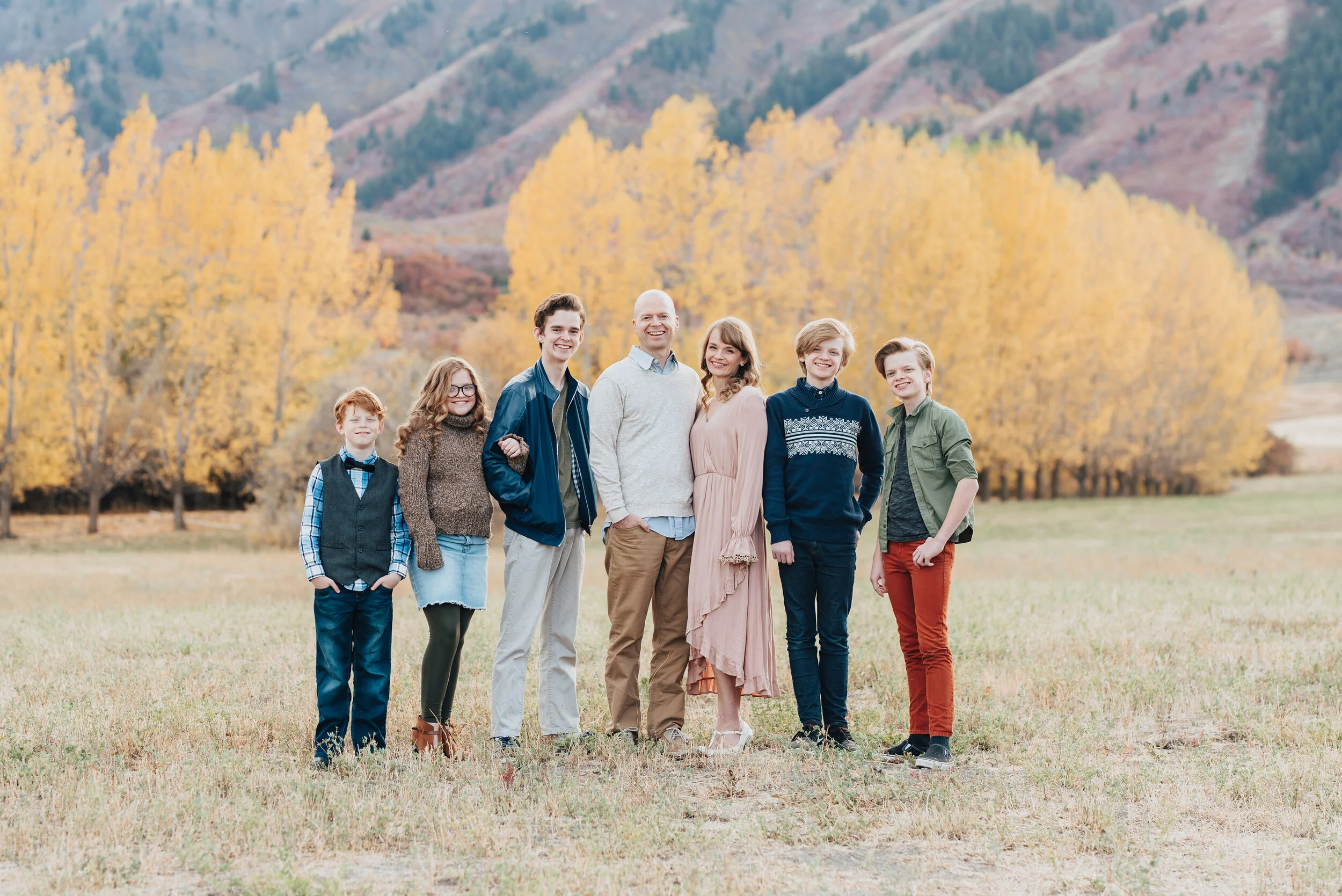 Outdoor family portrait in Logan, Utah featuring a family of seven, smiling and posed, dressed in Fall colors with yellow-leaved trees and mountains in the background, photographed by Kristi Alyse Photography. quality photography family and wedding …
