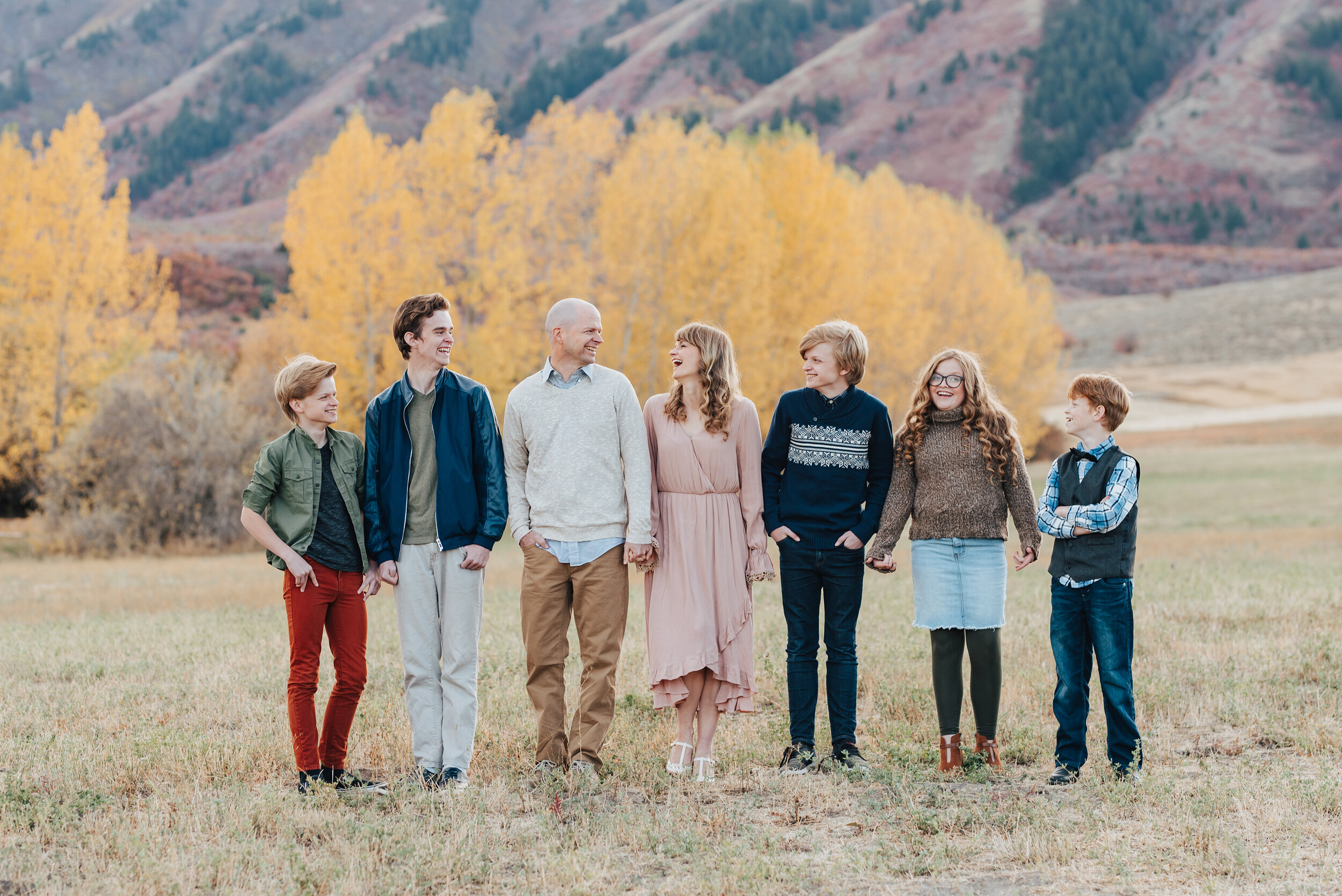 A candid outdoor family portrait in Logan, Utah featuring a family of seven smiling and laughing, dressed in Fall colors with yellow-leaved trees and mountains in the background, photographed by Kristi Alyse Photography. quality photography meaningf…