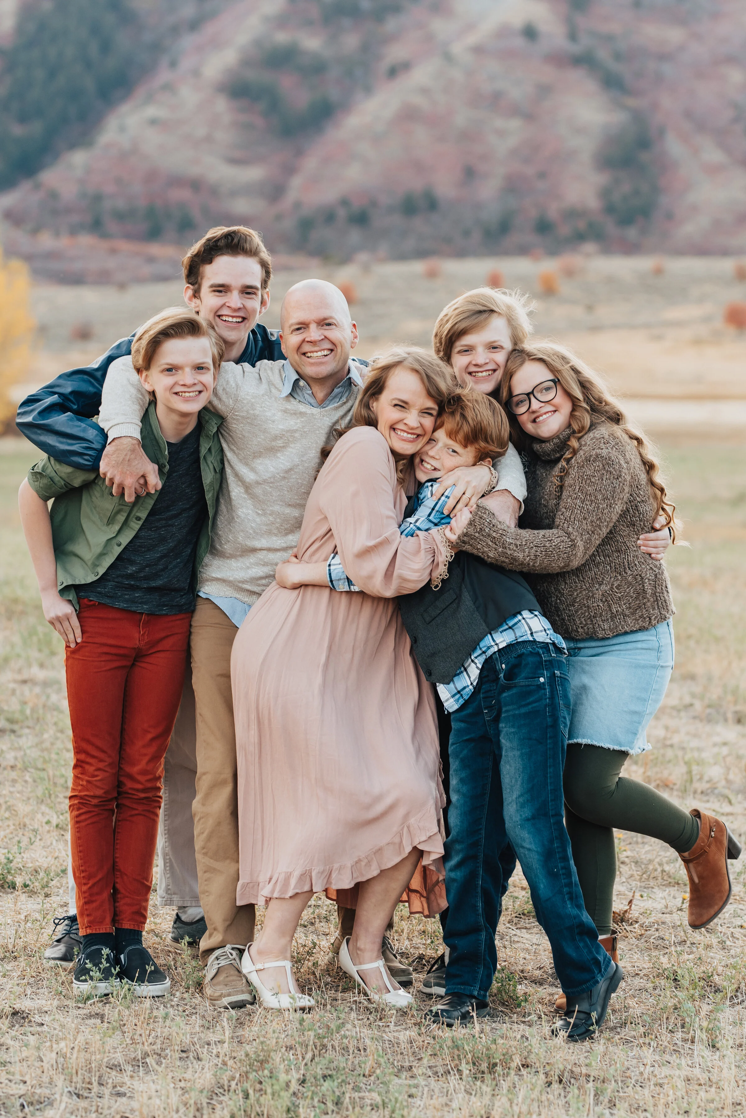  A candid outdoor family portrait in Logan, Utah featuring a family of seven smiling and hugging, dressed in Fall colors with trees and mountains in the background, photographed by Kristi Alyse Photography. quality photography meaningful family photo