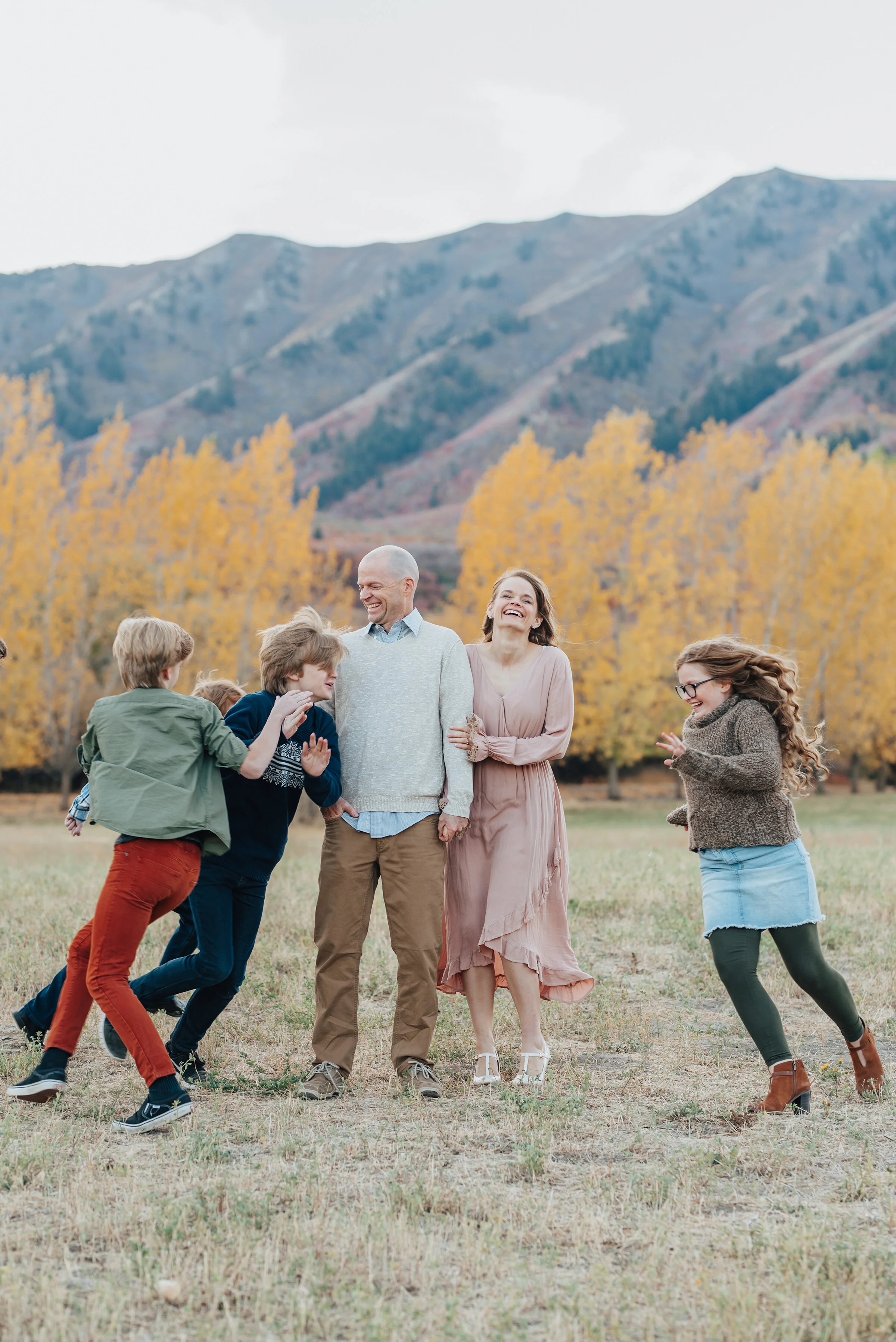  A candid family portrait in Logan, Utah featuring mom and dad standing still while their kids run around them, dressed in Fall colours with trees and mountains in the background, photographed by Kristi Alyse Photography. quality family photography U