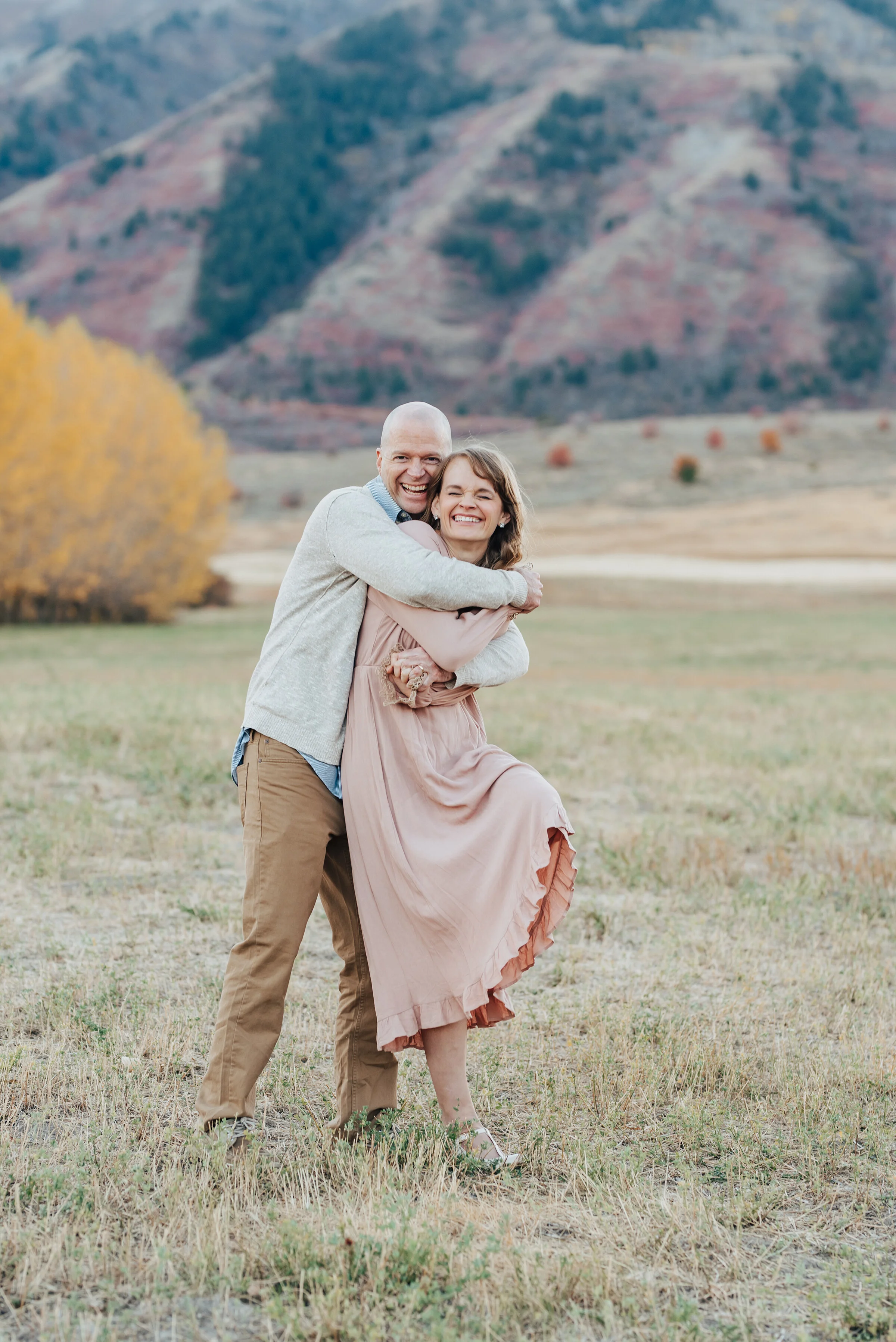  A candid outdoor family portrait featuring the dad and mom, hugging and smiling at the camera, with yellow trees and mountains in the background, photographed by Kristi Alyse Photography. quality wedding photography family photography meaningful fam