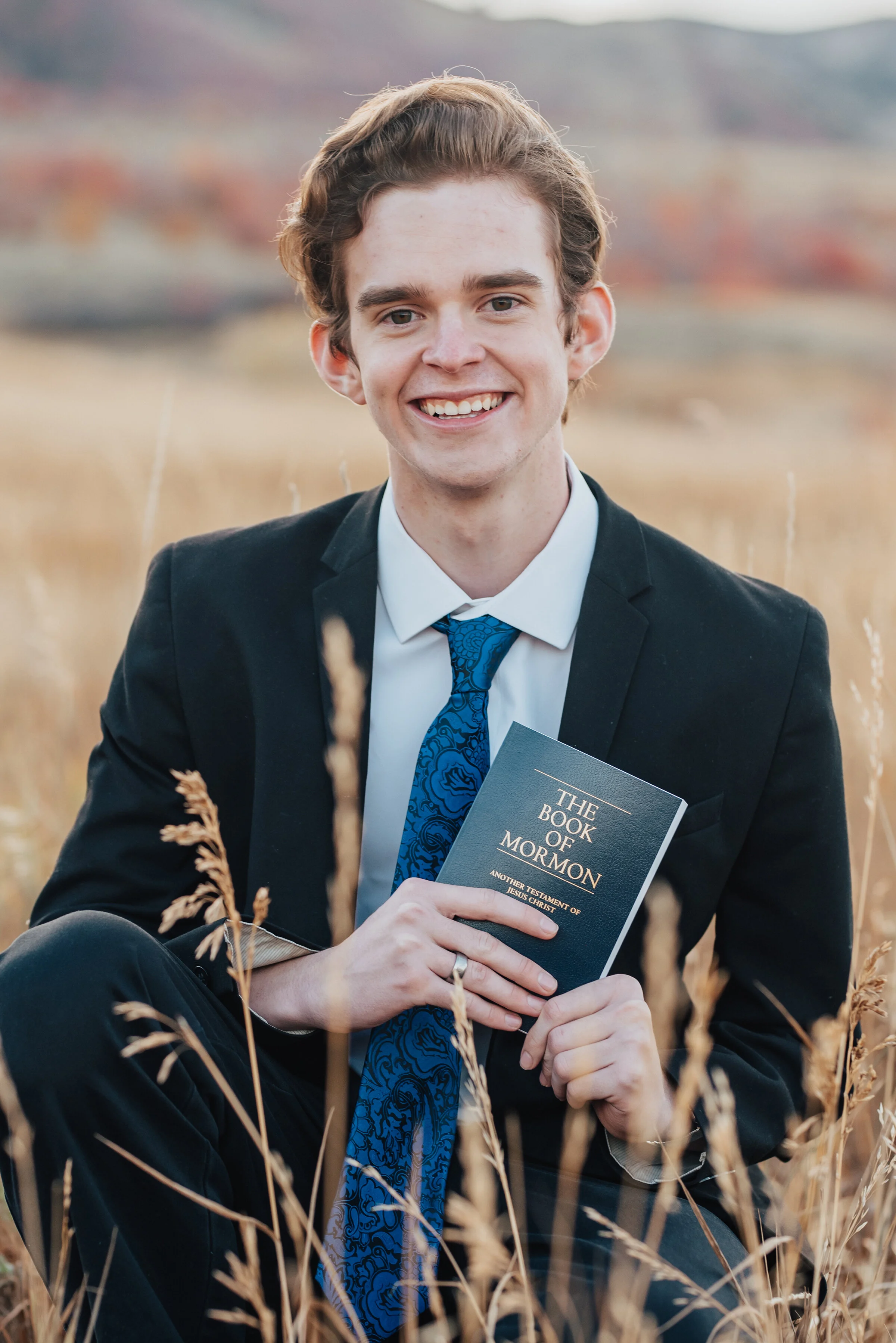  Close-up portrait of a young man, crouching in a field, dressed in a dark suit, white shirt, and blue tie, holding a copy of the book of Mormon, photographed by Kristi Alyse Photography. quality wedding and family photography meaningful family photo