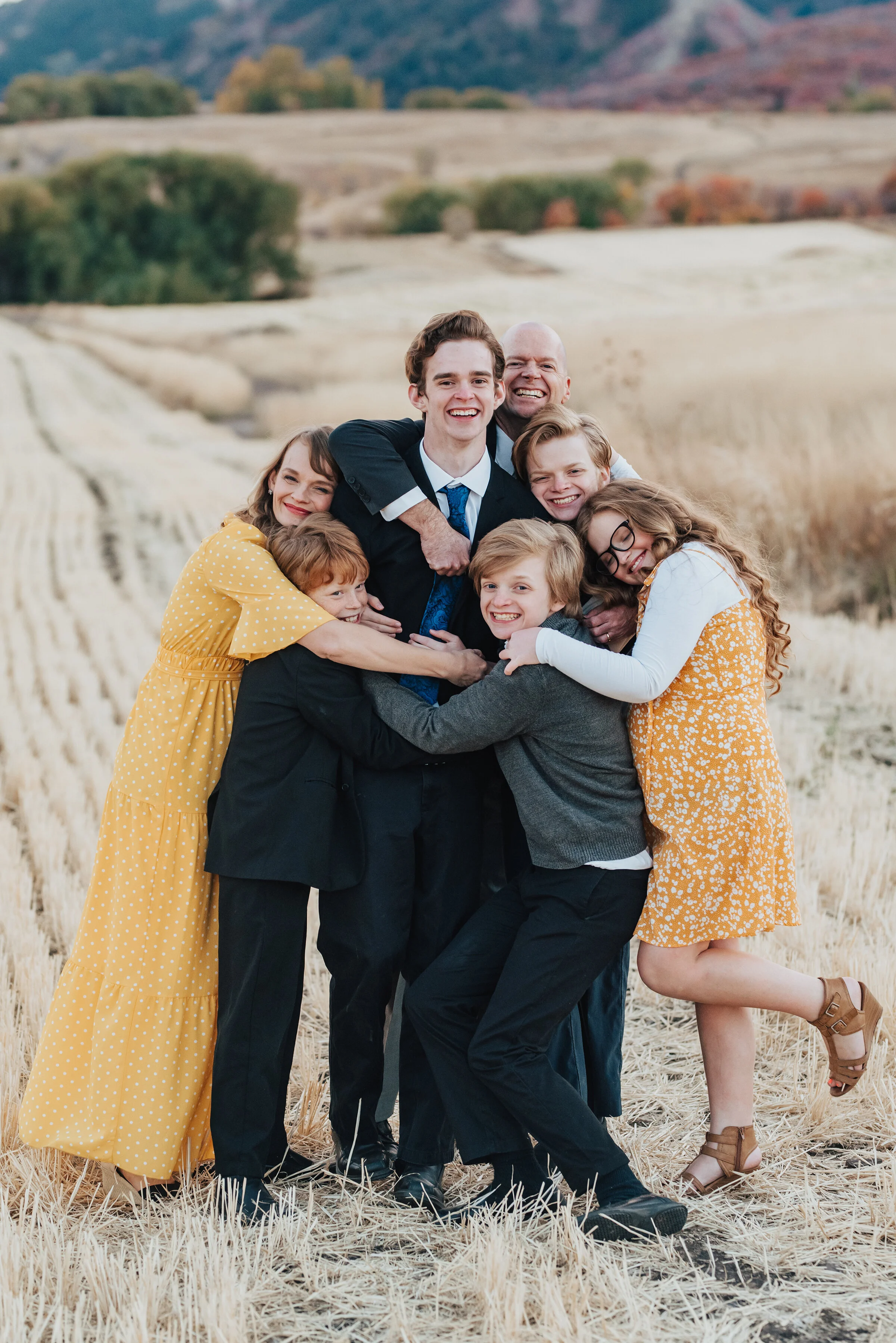  A portrait of a family of seven, hugging each other in an empty farm field, males dressed in dark colours, the women dressed in golden yellow, photographed by Kristi Alyse Photography. quality wedding and family photography meaningful family photogr