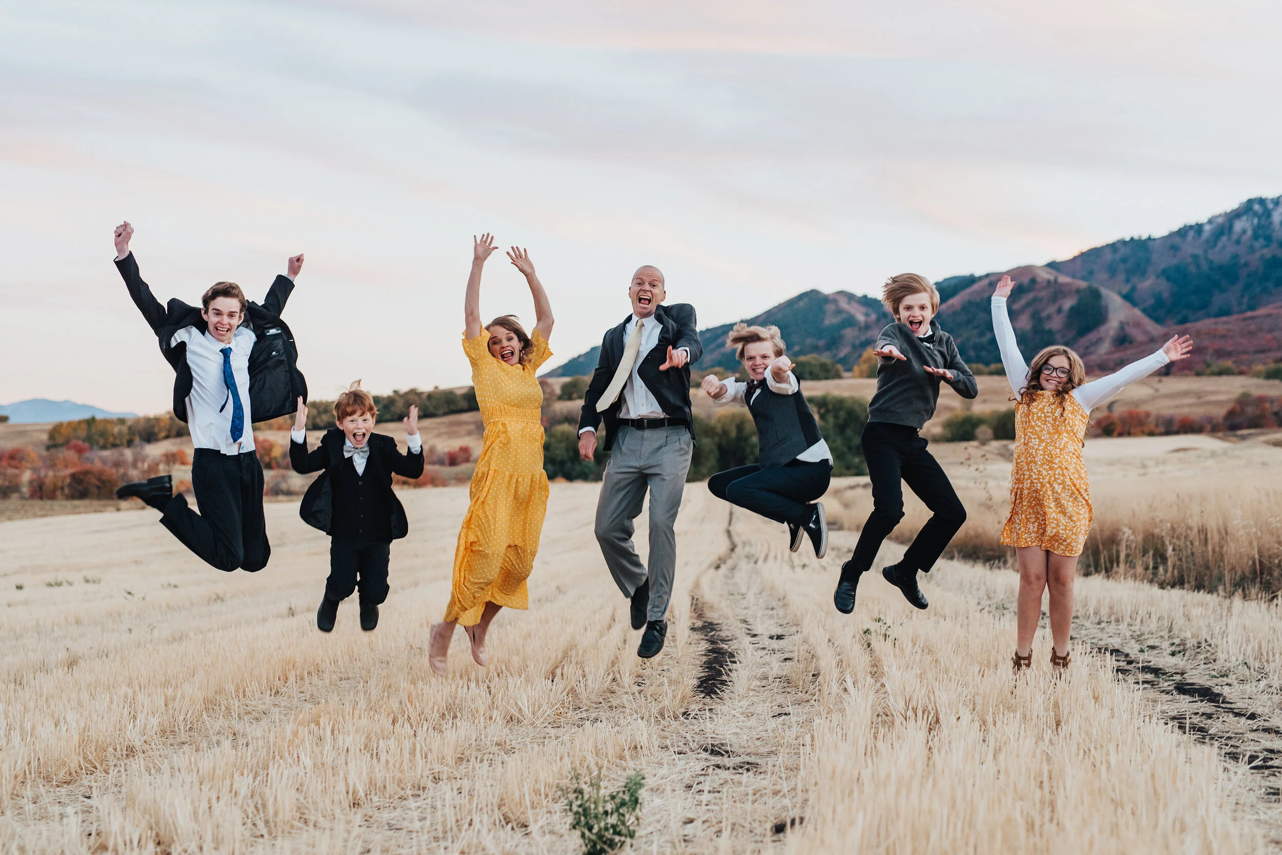 A family of seven jumping in the air, an empty farm field around them, boys dressed in dark colours, the girls dressed in golden yellow, photographed by Kristi Alyse Photography. quality photography wedding and family photography fun family photogra…