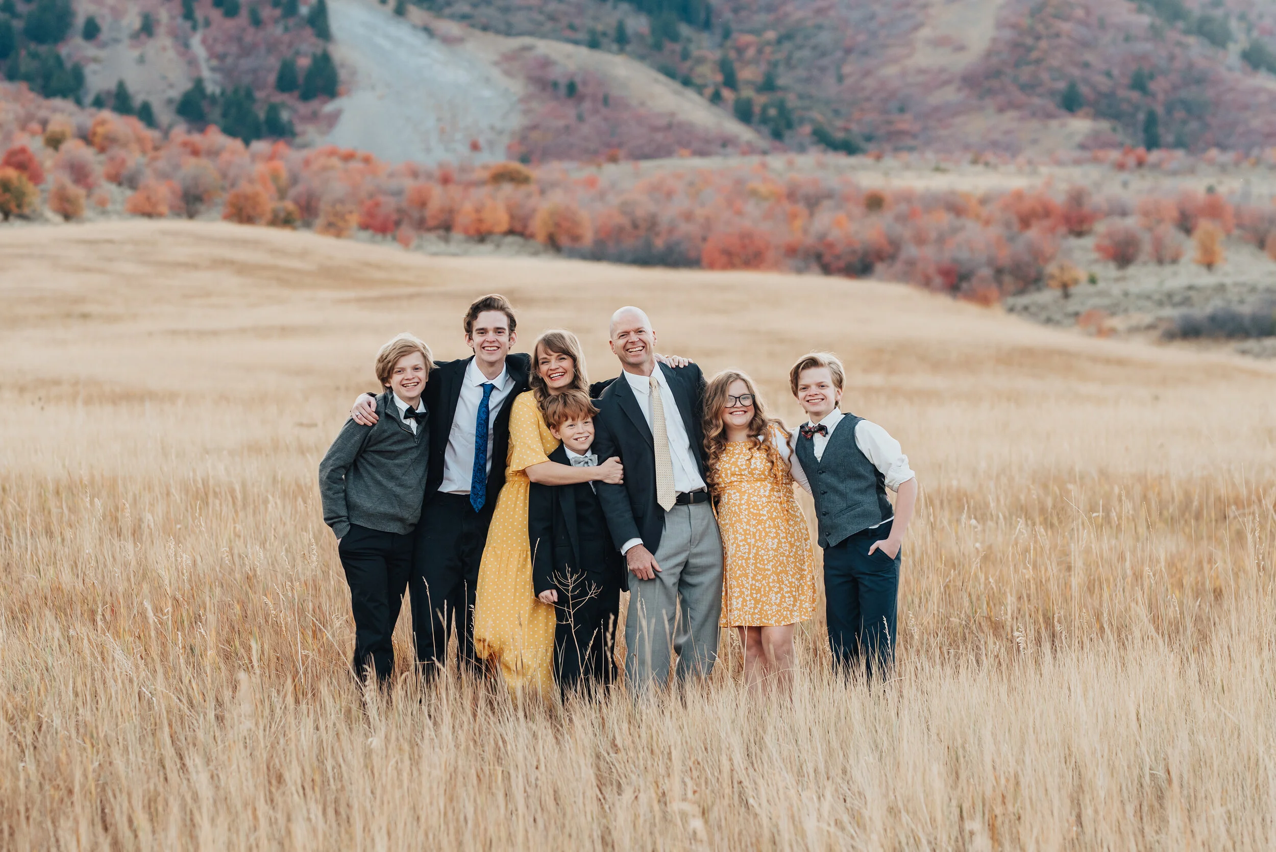  Portrait of a family, arms around each other, smiling at camera, an open field and autumn trees in the background, the males in dark suits with white shirts while the females are in golden yellow dresses, photographed by Kristi Alyse Photography. qu