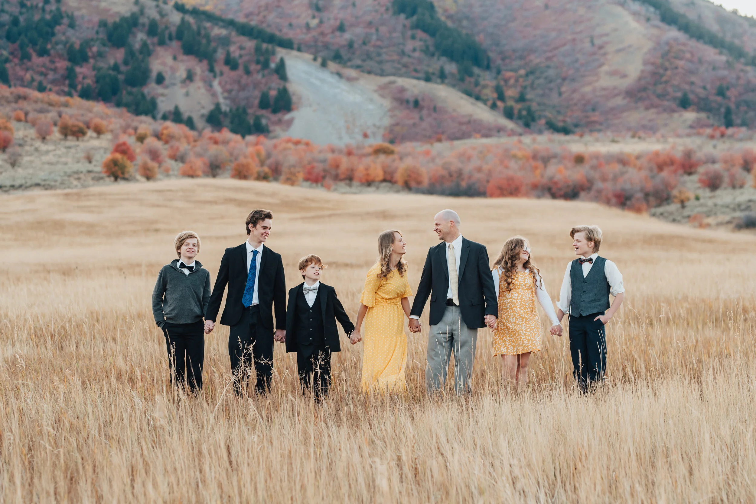  A family of seven, standing in a row holding hands in a field of wild grasses, autumn trees in the background, the boys in dark suits with white shirts, the girls are in golden yellow, photographed by Kristi Alyse Photography. quality family photogr