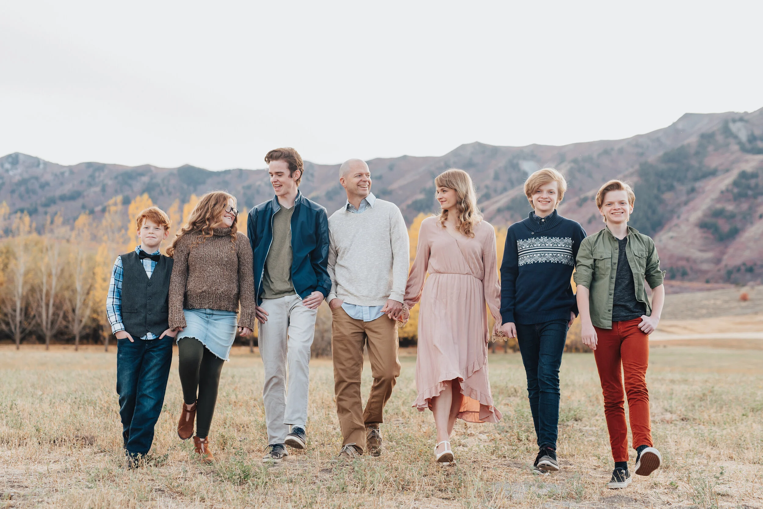 Outdoor family portrait in Logan, Utah featuring a family of seven, smiling and walking, dressed in Fall colors with trees and mountains in the background, photographed by Kristi Alyse Photography. quality wedding photography meaningful family photo…