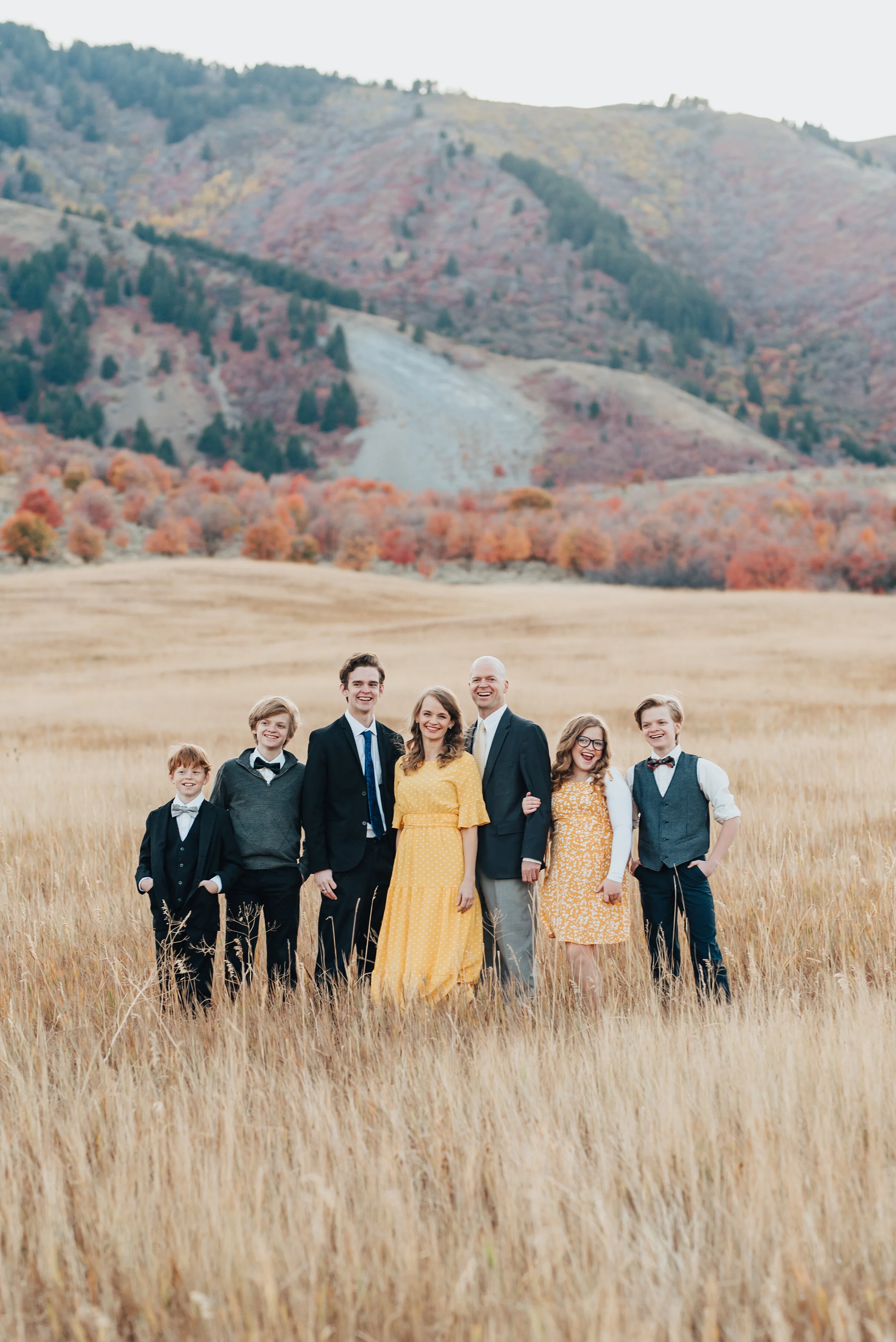  A family portrait in an open field, autumn trees and a mountain in the background, the males in dark suits with white shirts while the females are in golden yellow dresses, photographed by Kristi Alyse Photography. quality family photography meaning