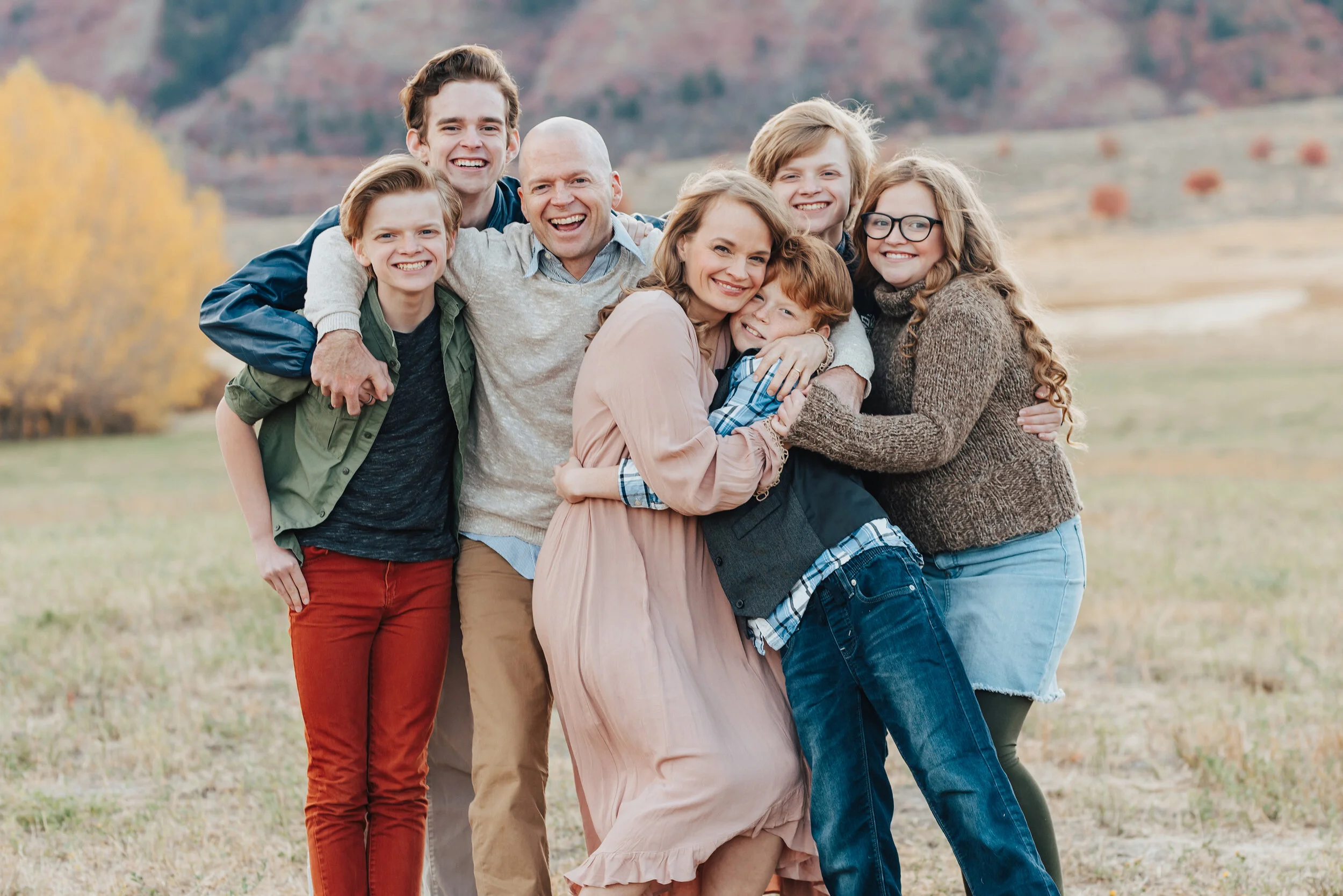 A portrait of a family of seven smiling and hugging, looking at the camera, dressed in fall colours, with an open field and trees in the background, photographed by Kristi Alyse Photography. quality family photography meaningful family photography L…