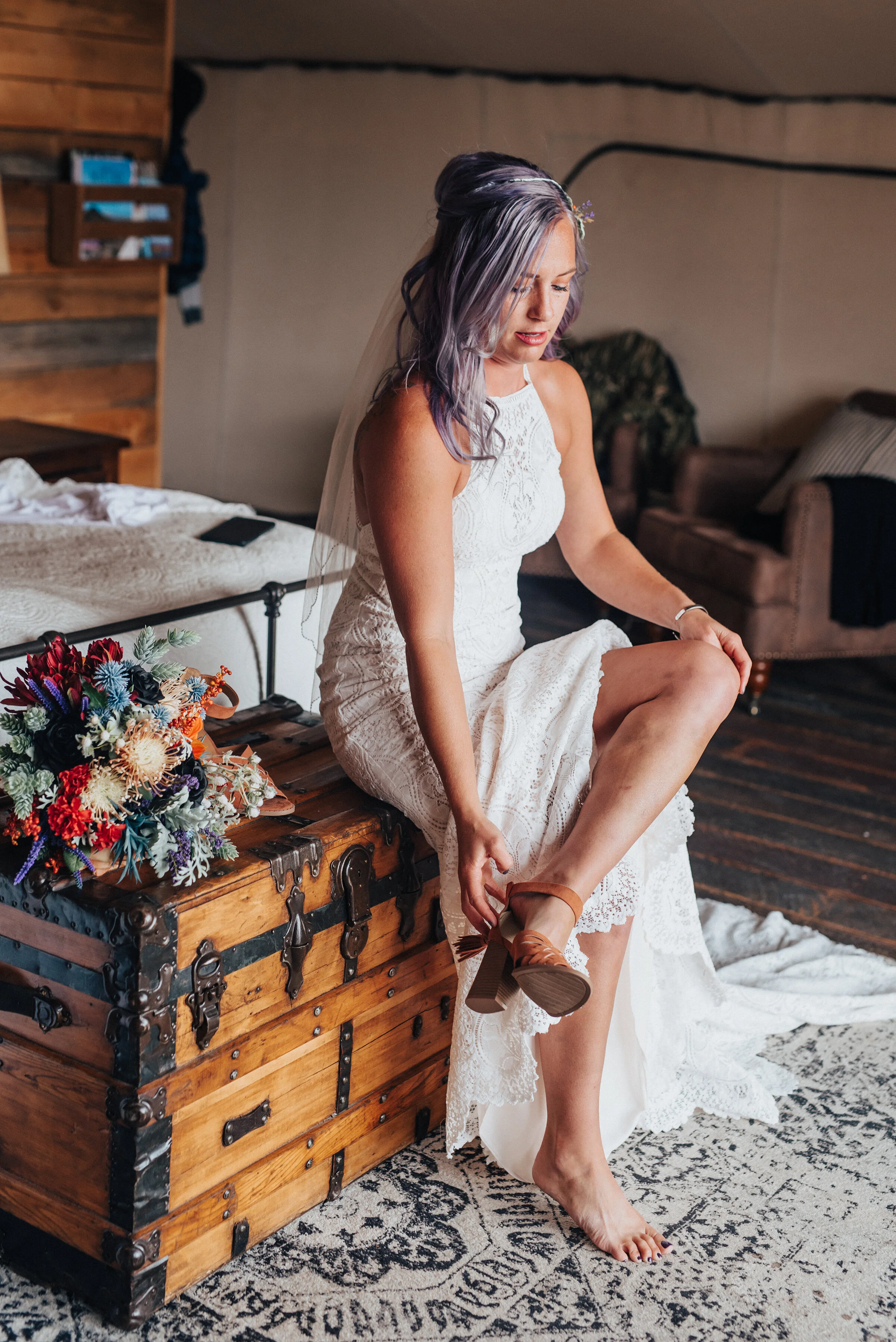  Boho bride with lace dress and purple and orange flower bouquet sitting on vintage chest and boho rug in this candid photo in Logan Utah by Kristi Alyse Photography. meaningful photography northern utah wedding and family photographer quality photog