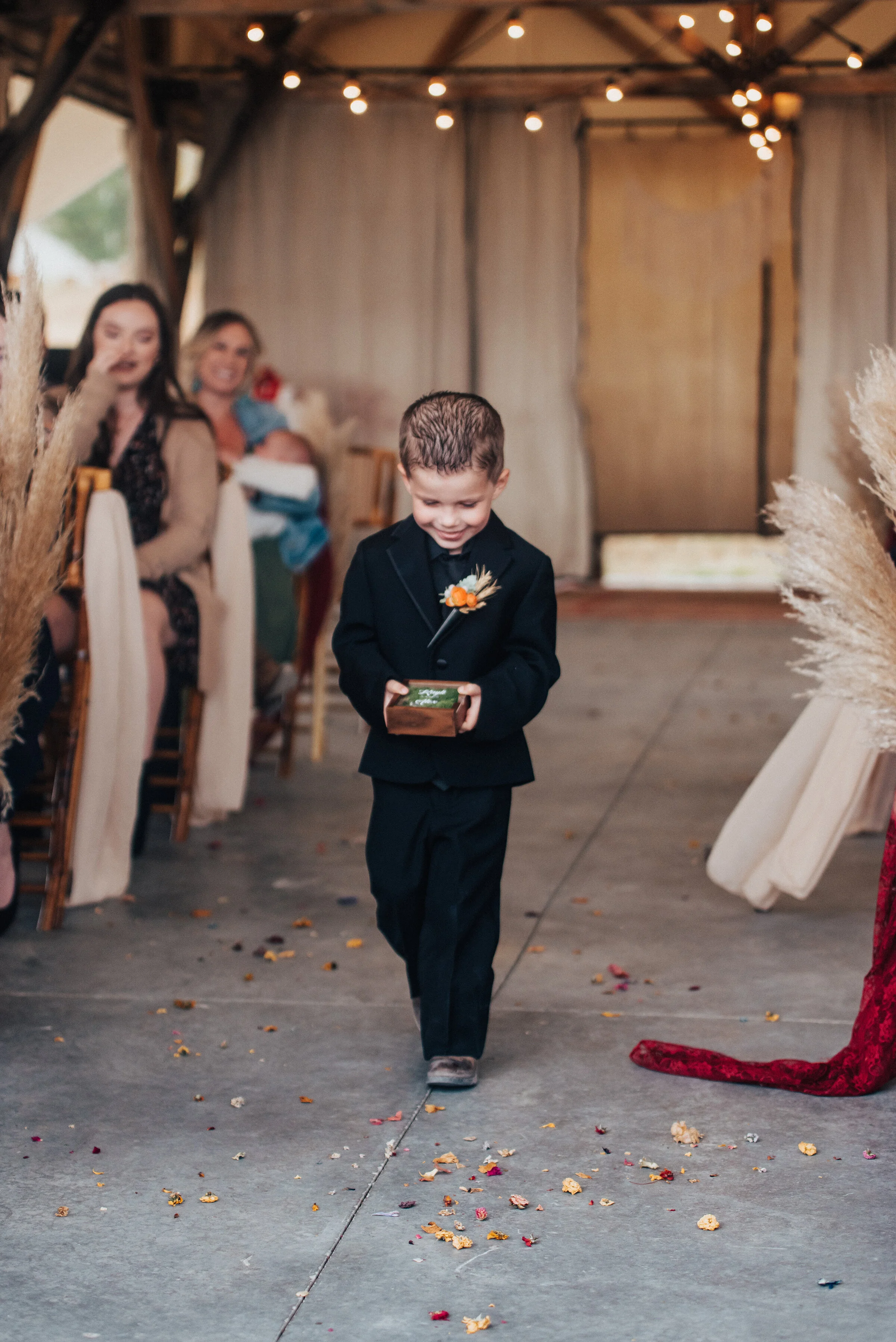  Adorable groomsmen boy in black suit walking down the aisle with string lights and fall boho decor in this candid photo in Northern Utah by Kristi Alyse Photography. logan utah wedding and family photographer boho wedding groomsmen child wedding ais