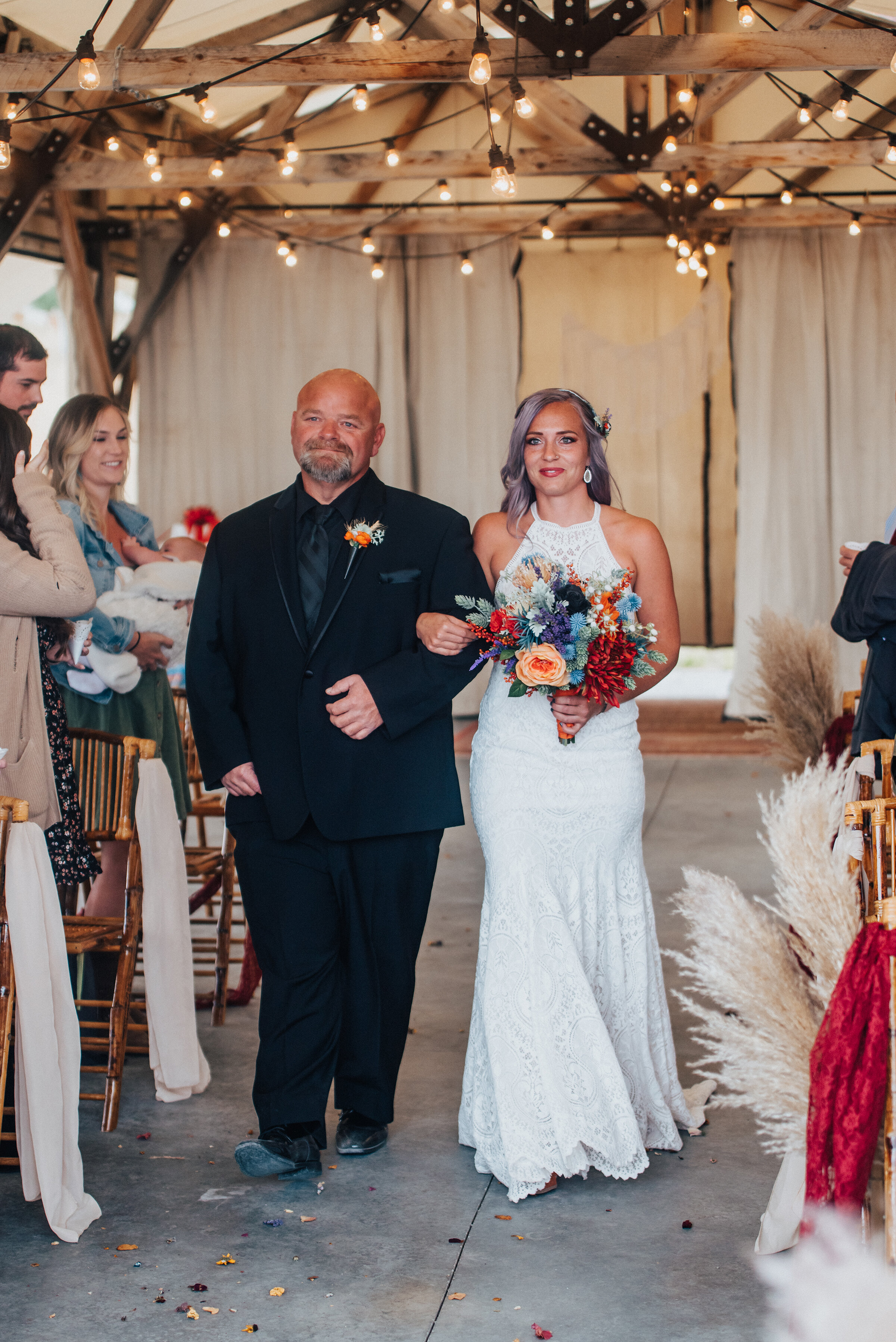  Beautiful bride and father walking down the wedding aisle with string lights and boho fall wedding decor in this candid photo by Kristi Alyse Photography in Logan Utah. northern utah black suit orange and purple flowers lace wedding dress wedding ai