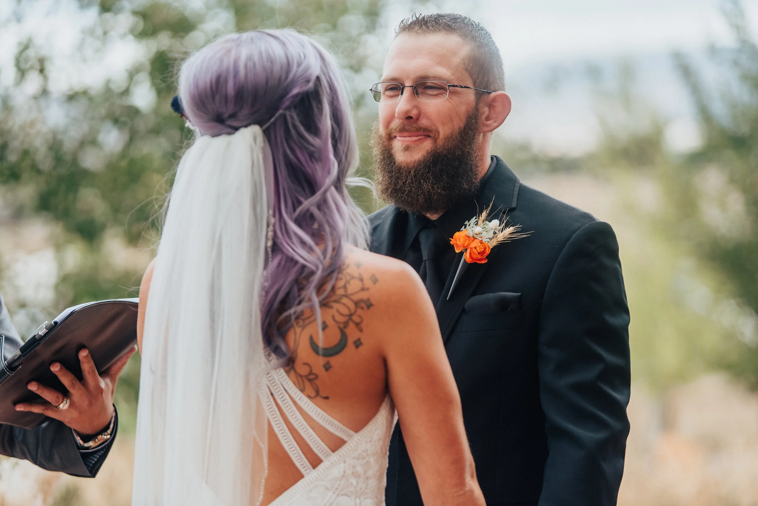 Groom in black suit and orange boutonniere and bride with purple hair and veil and open back lace dress in this candid photo in Logan Utah by Kristi Alyse Photography.&nbsp; Northern utah boho wedding lace open back purple hair orange flowers black …
