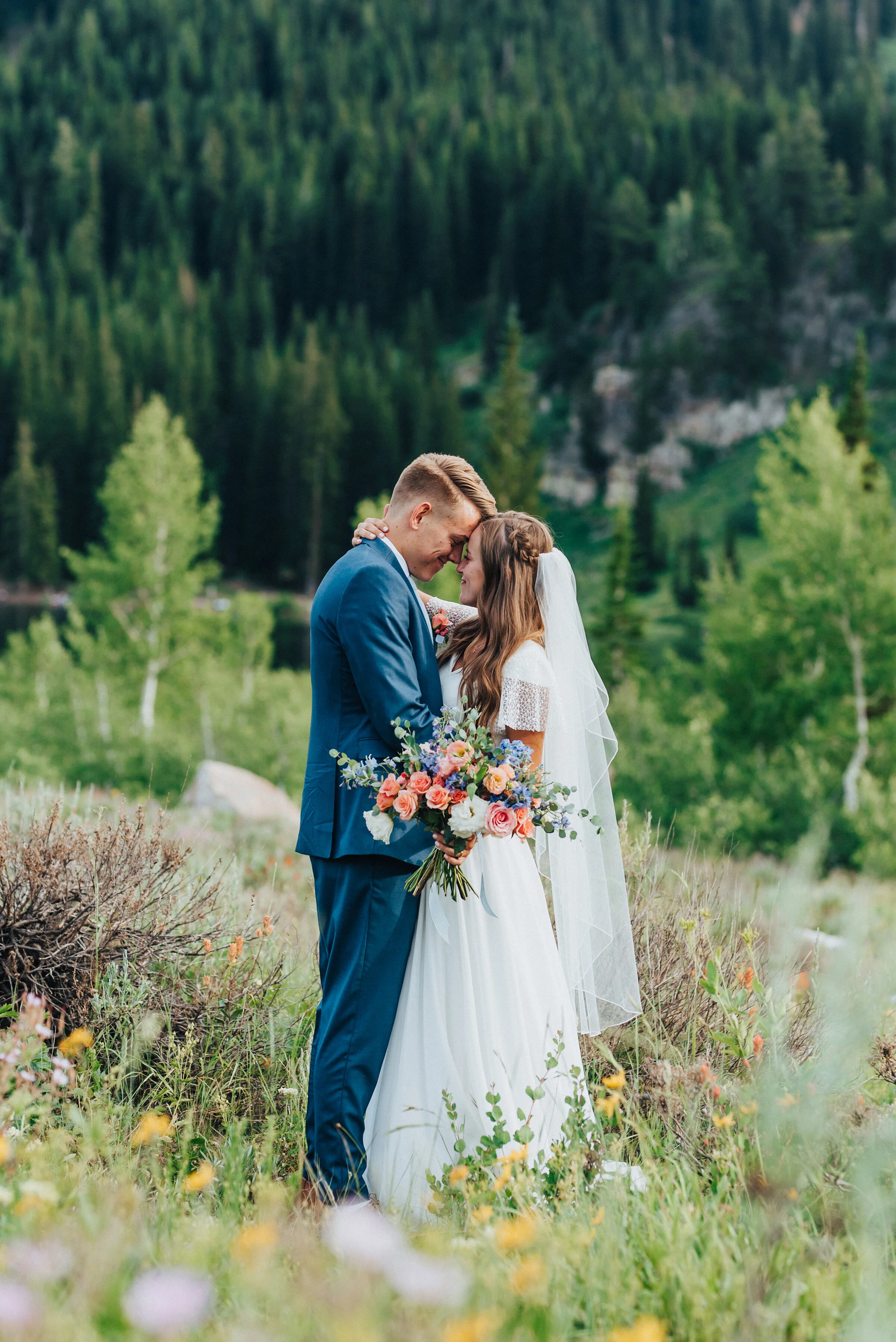  Gorgeous bride in a modest wedding dress and mid-length wedding veil being kissed by her groom in a blue suit at Tony Grove while holding a bright wildflower bridal bouquet. Tony grove wedding formals northern utah wedding photography bridal bouquet
