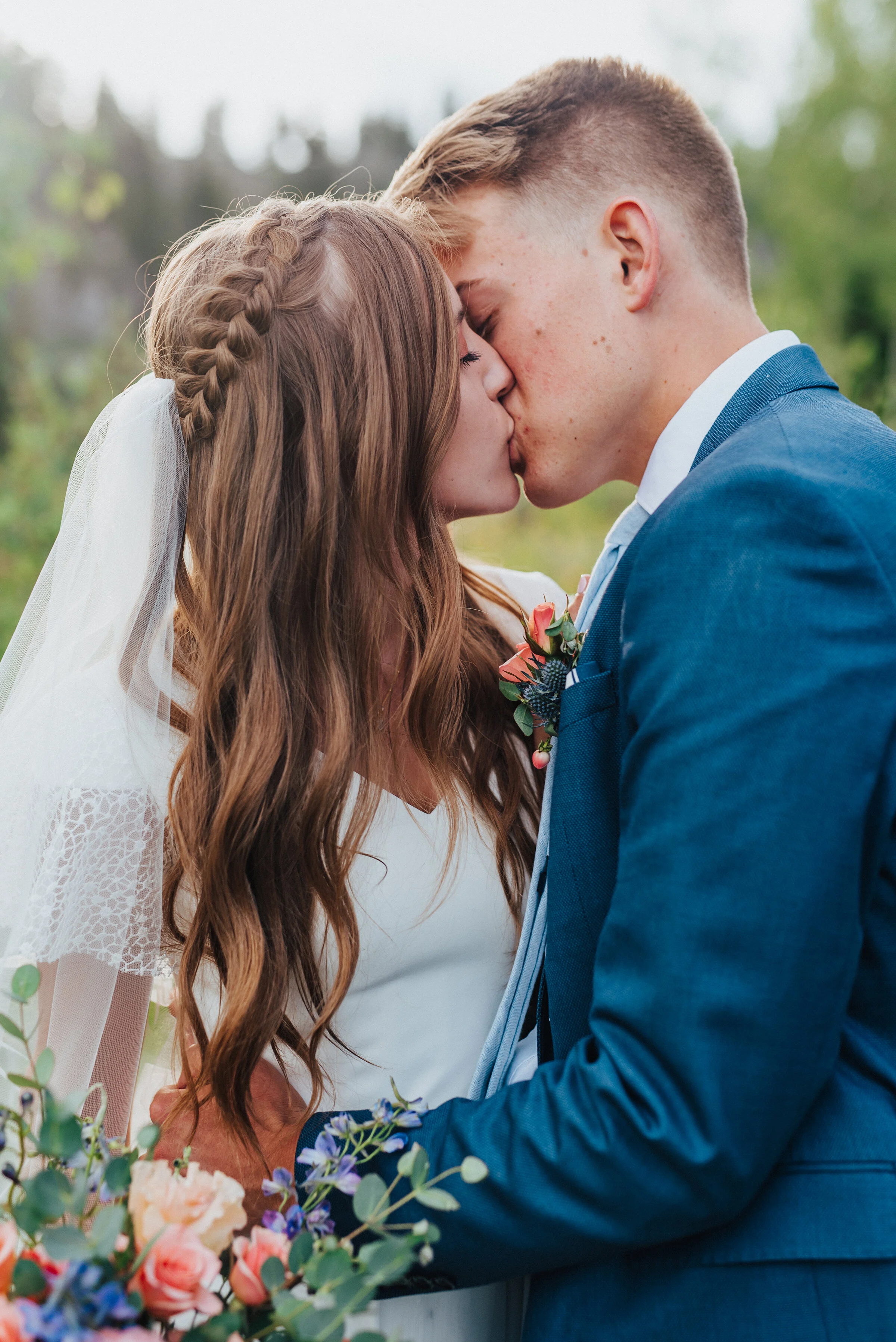 Close-up shot of a bride and groom kissing accenting her cascading hair with the veil tucked into her braid while holding her bridal bouquet. Northern utah wedding photography boutineer for the groom bridal bouquet inspo wedding veil hair and makeup