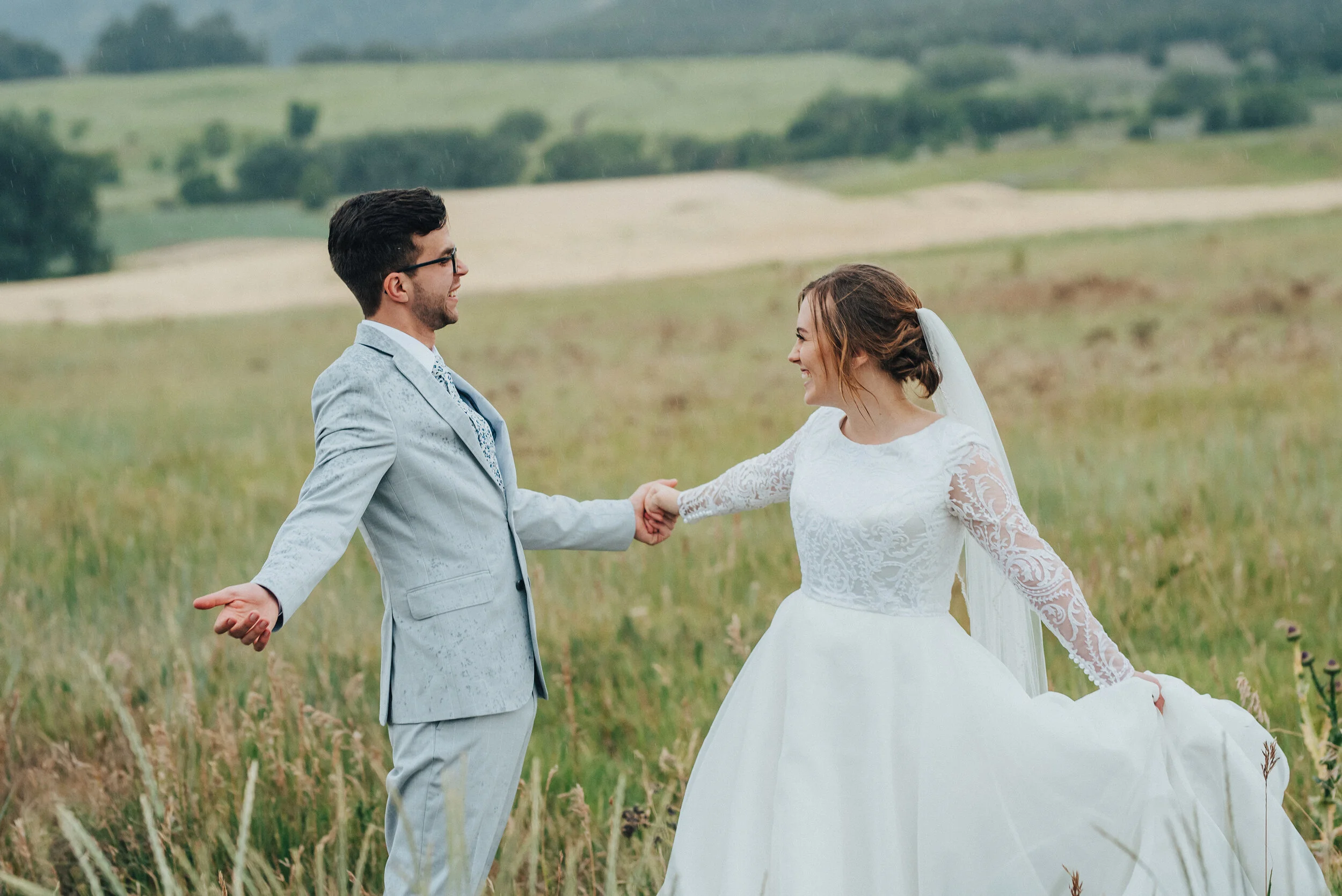 Dancing in the rain! A sunny bridal session turned sudden downpour made for a playful pictures captured by Kristi Alyse Photography in Cache Valley, Utah. Laced long sleeved wedding dress, twill gray wedding suit, floral groom’s tie, rainy bridal se…