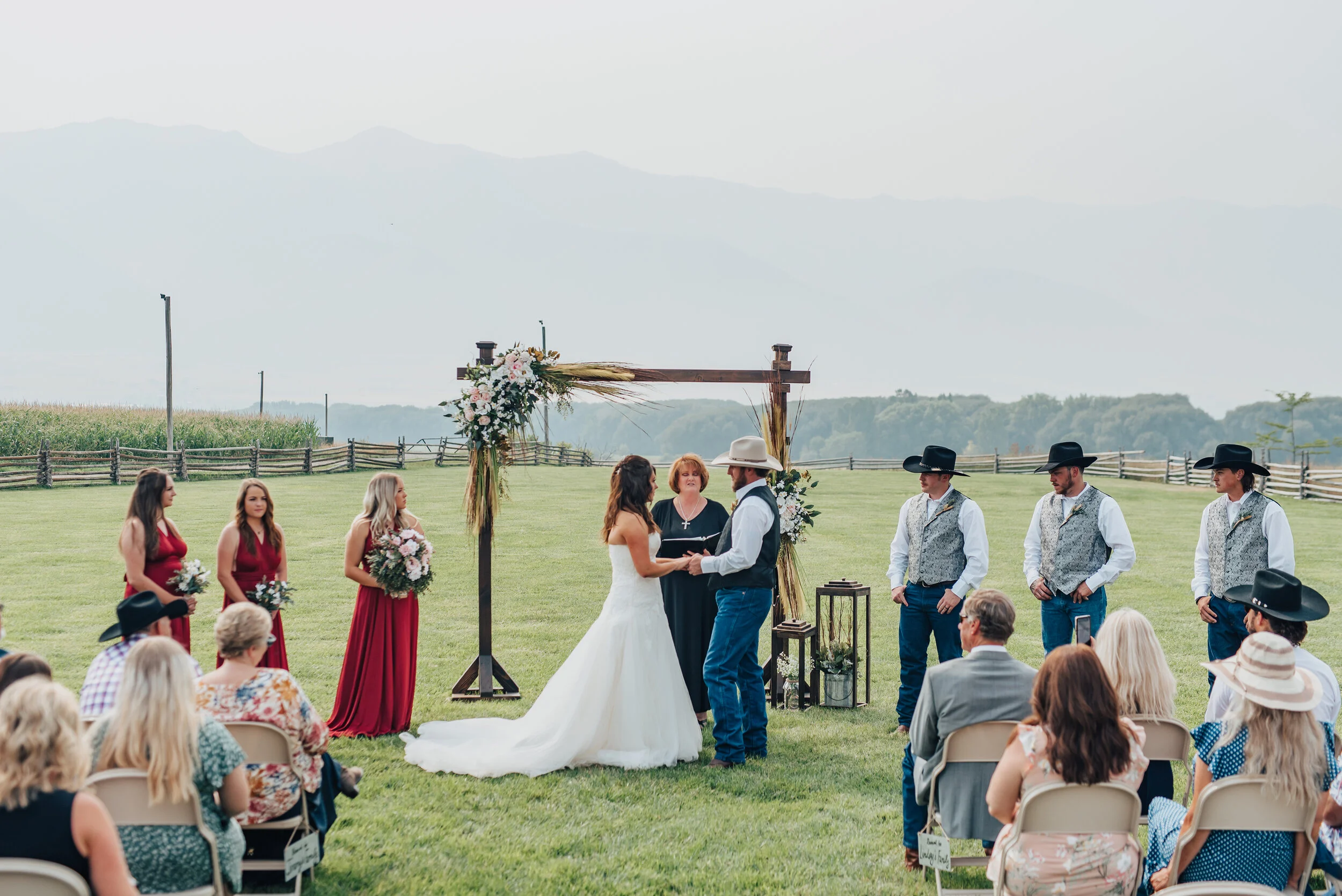 Bride and Groom lovingly hold hands beneath a simplistic wooden wedding arch during their ranch wedding ceremony captured by Kristi Alyse Photography in Logan, Utah. long ruby red bridesmaids dresses, wooden wedding arch, ranch wedding, outdoor wedd…