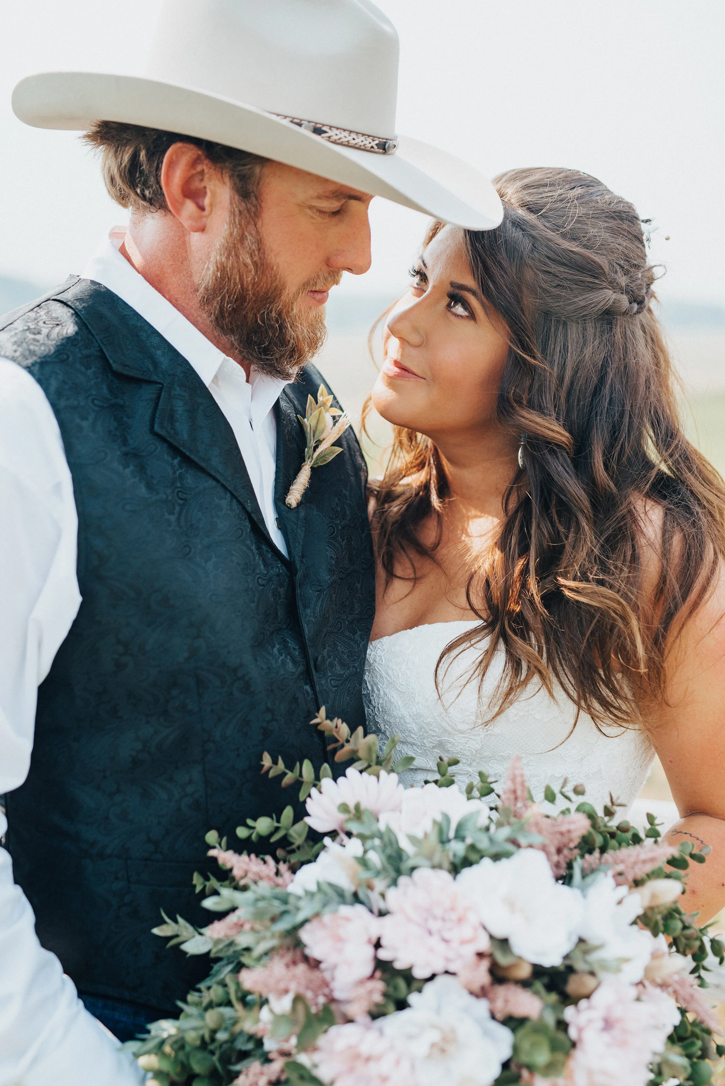  Southern bride and groom fondly embrace one another during their wedding portraits shot by Kristi Alyse Photography in Wellsville, Utah. White colored wedding cowboy hat, white shirt and black vest wedding clothes, blush pink and white weding bouque