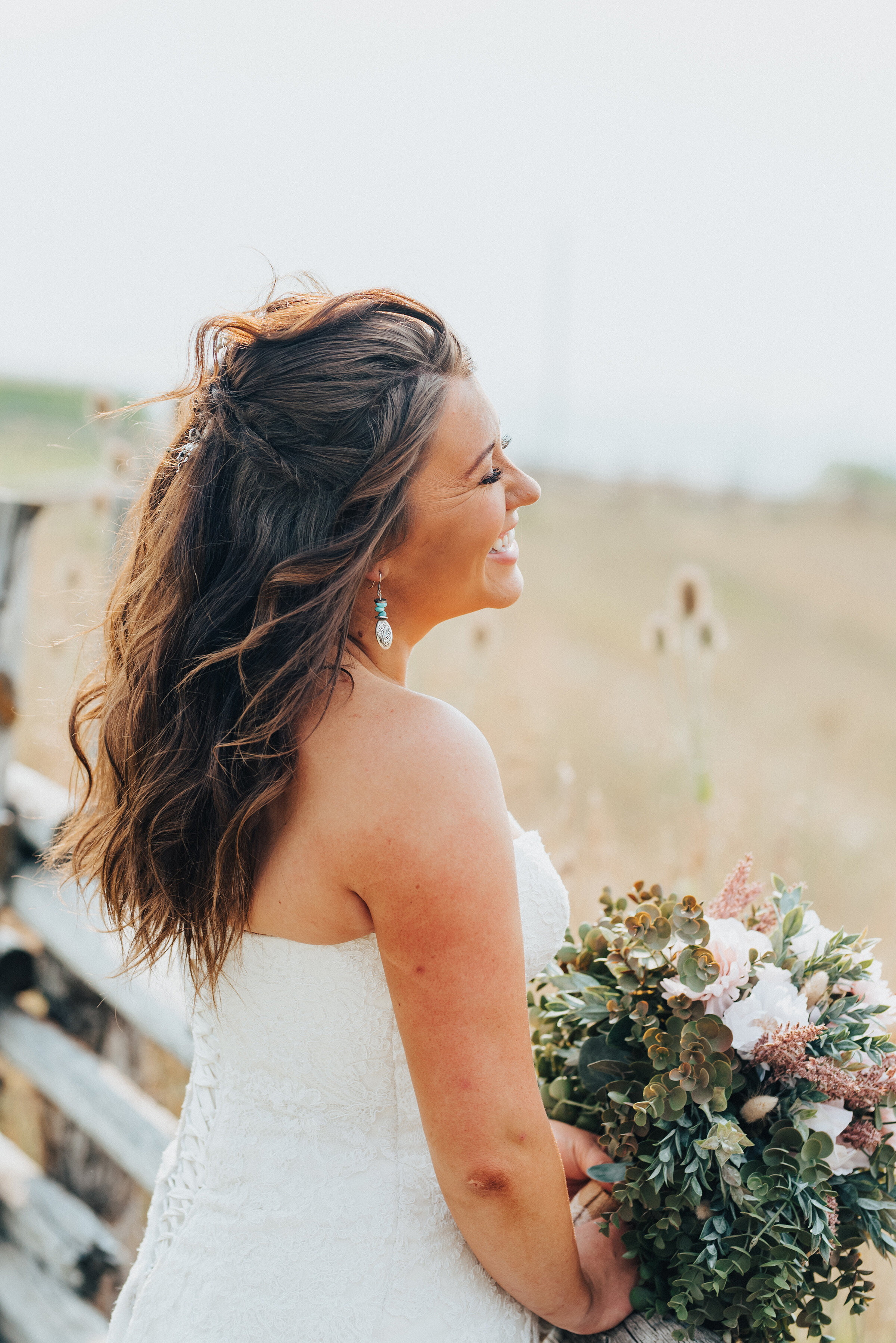  A beaming bride overlooks vast Utah fields while posing for her wedding portraits taken by Kristi Alyse Photography in Wellsville, Utah. barn wedding, medium length messy curled wedding hair, southern wedding, lace up strapless wedding dress, wooden
