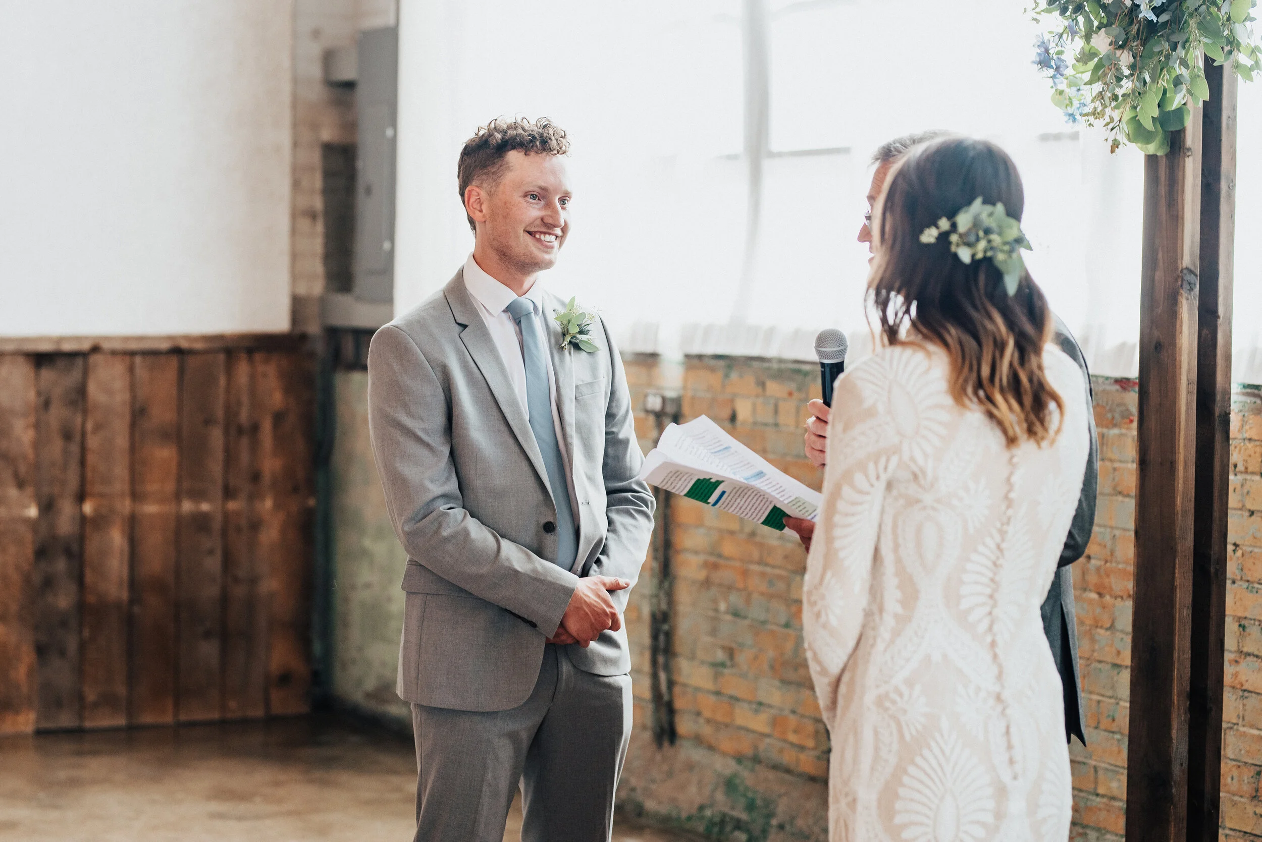  Kristi Alyse Photography captures bride and groom exchanging wedding vows as the sun peeks through the large industrial windows of the Bright Building in Provo, Utah. Bright building provo utah, wedding vows, loose curled half wedding updo, button d