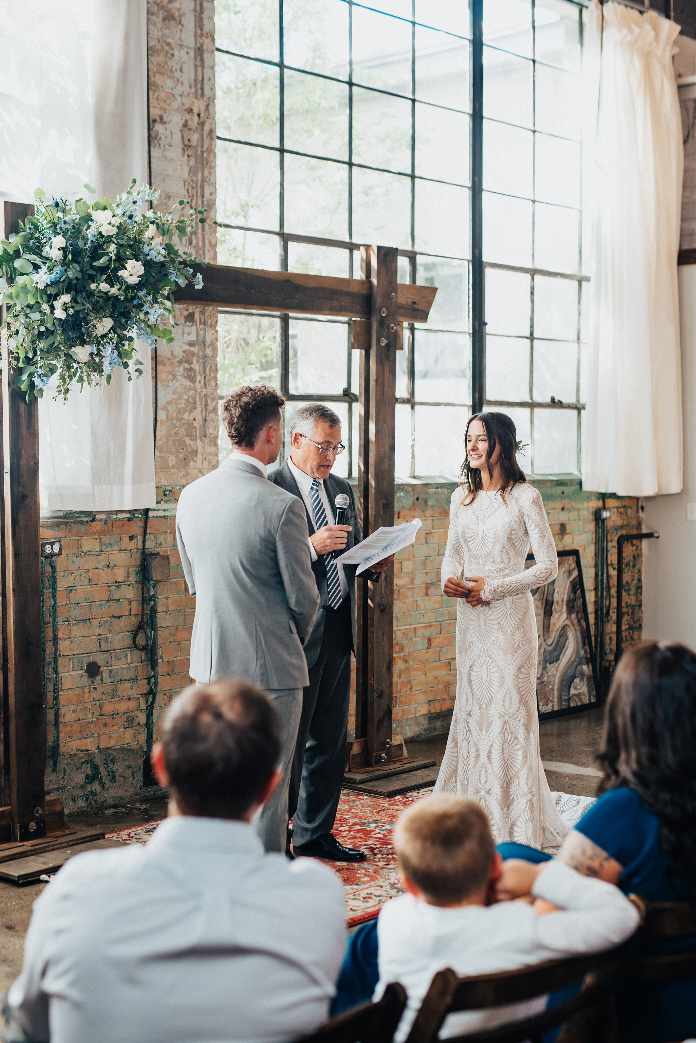 Kristi Alyse Photography photographs the quiet light peeking through the large industrial windows of the Bright Building, as an intimate wedding ceremony commences in Provo, Utah. bohemian styled wedding ceremony, vintage rug, wooden wedding ceremon…