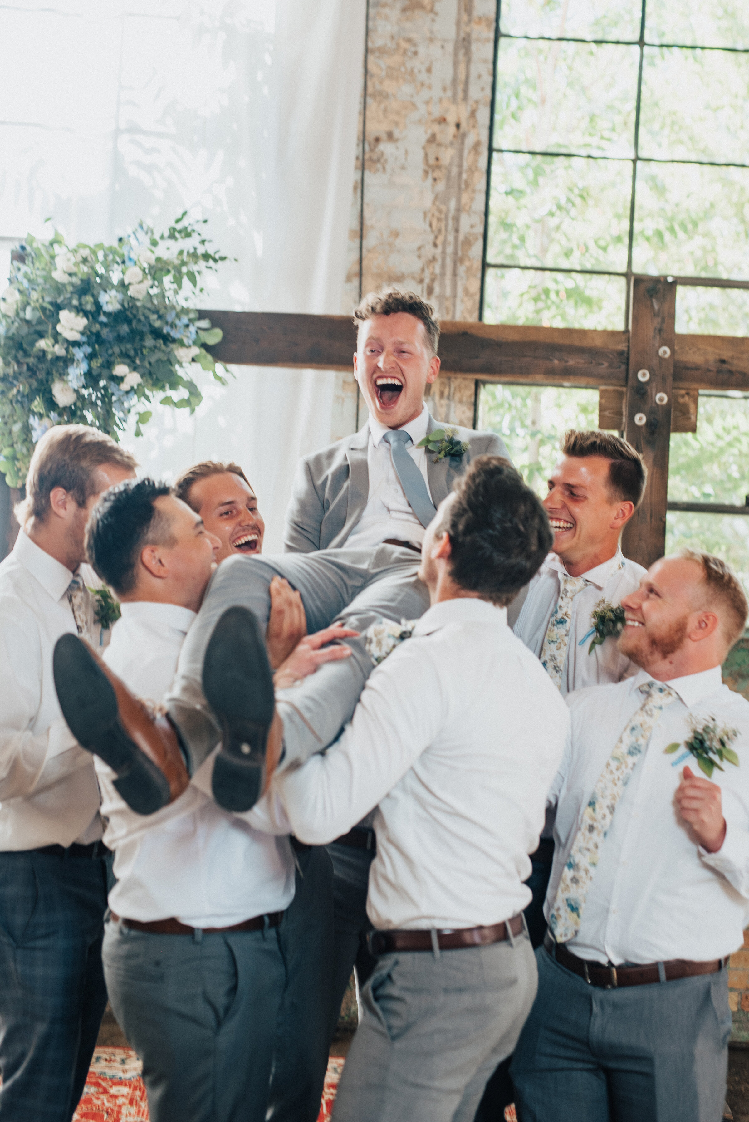  Kristi Alyse Photography captures the bustle and energy of a newly wed groom being tossed in the air by his groomsmen at the Bright Building in Provo, Utah. groomsmen toss, gray groomsmen pant, gray wedding suit, blue gray wedding tie, industrial pr