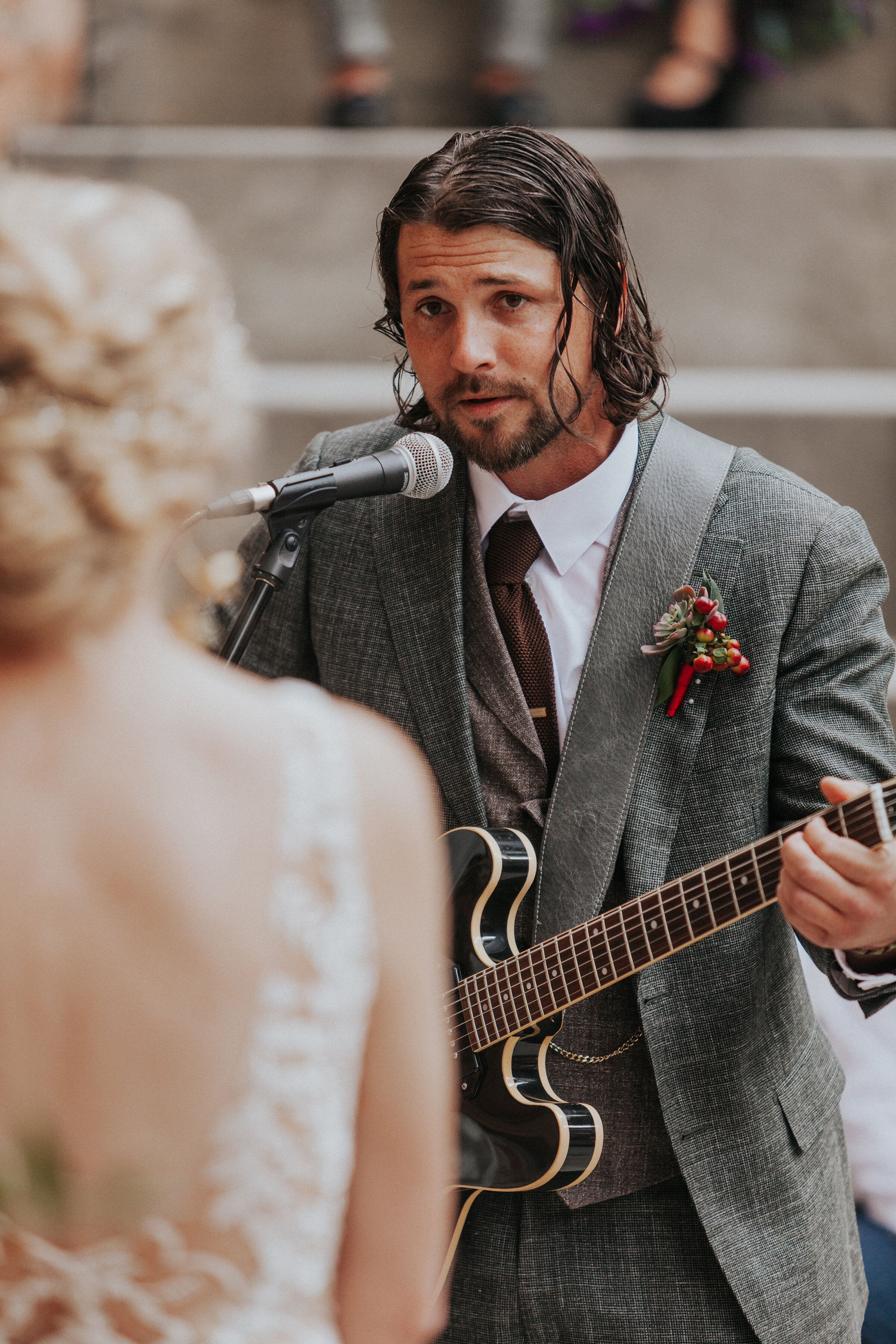 Handsome groom with long curly hair and facial hair wearing a dark gray suit playing the guitar for his bride during the wedding ceremony by Kristi Alyse Photography in Northern Utah. ideas for ceremony boho wedding ceremony groom outfit inspo for b…