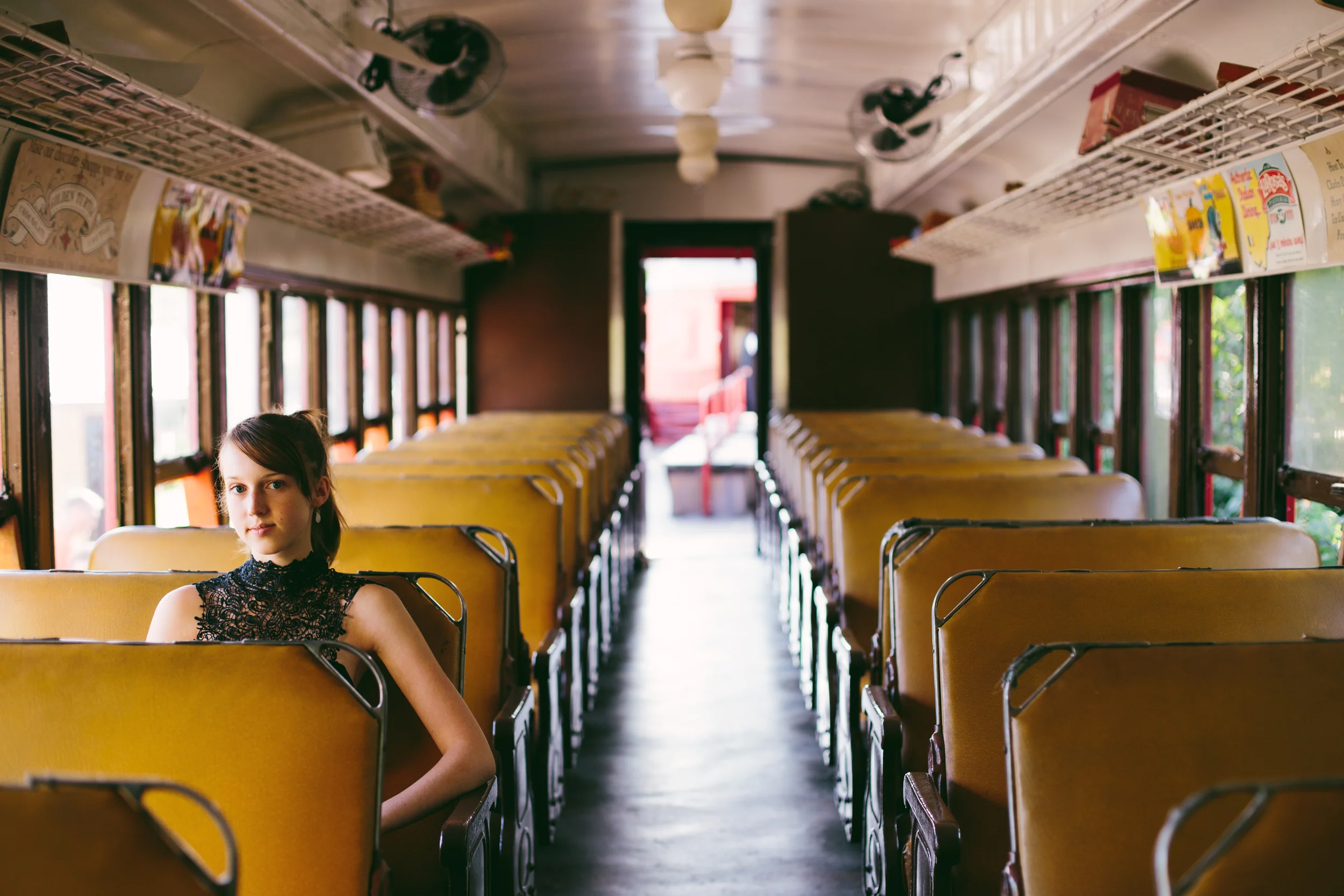 Inside a vintage school bus with yellow seats and large windows, a young woman with dark hair in a ponytail wearing a black lace top is seated on the left side, looking directly at the camera. The bus's interior features overhead wire racks, some wit