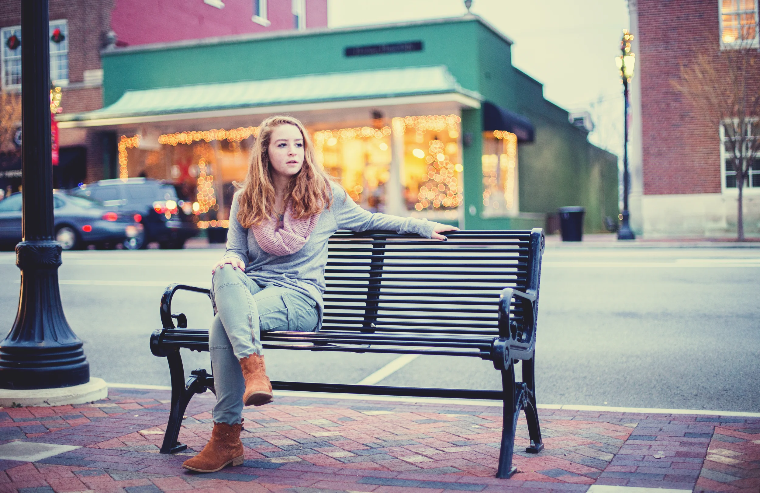 A young woman with red hair and freckles sitting on a black park bench on a city sidewalk, wearing a gray sweater, pink scarf, jeans, and brown boots, with a store and parked cars in the background during dusk.