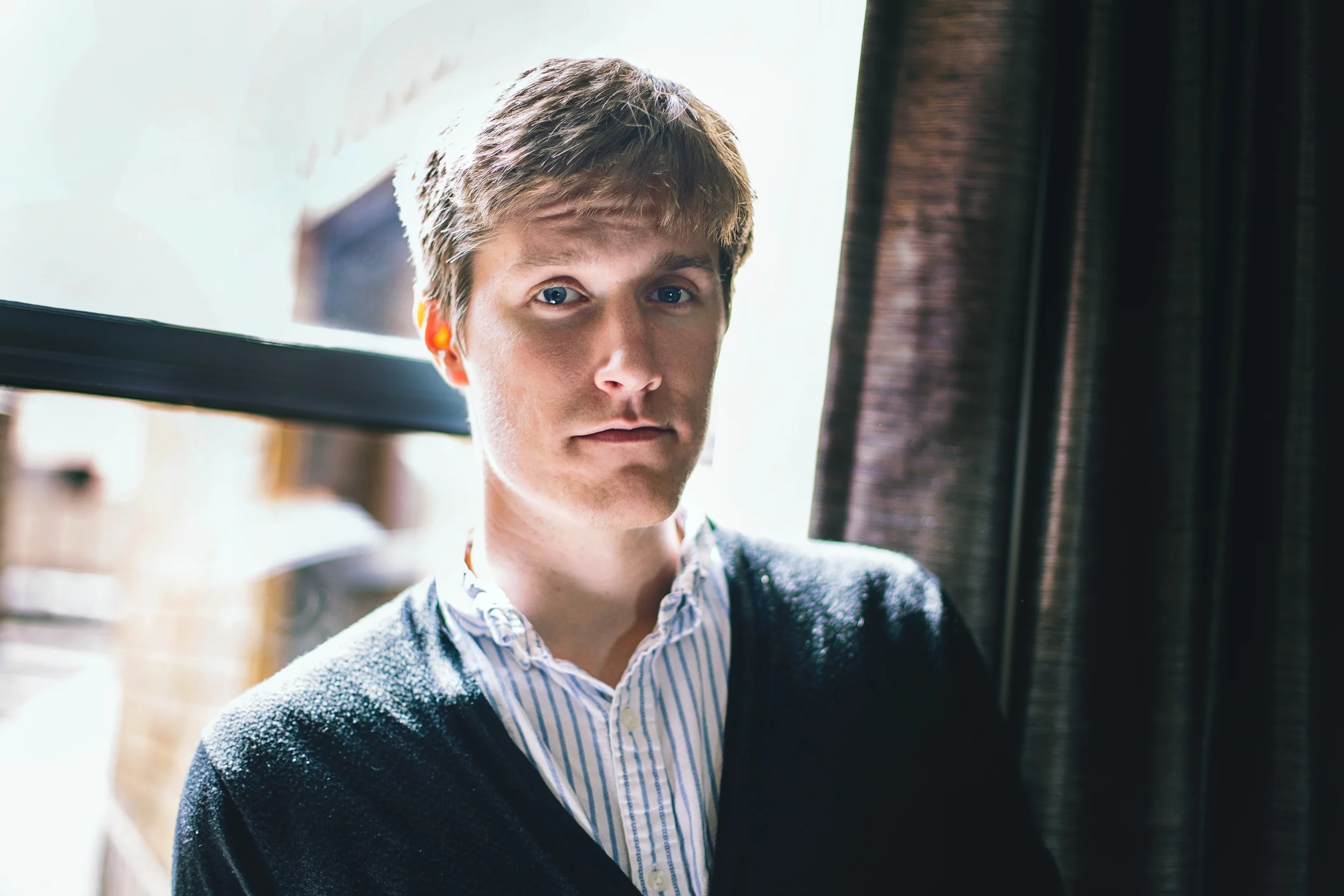 A young man with light brown hair and blue eyes looking at the camera, wearing a patterned shirt and a dark sweater, standing near a window with curtain.
