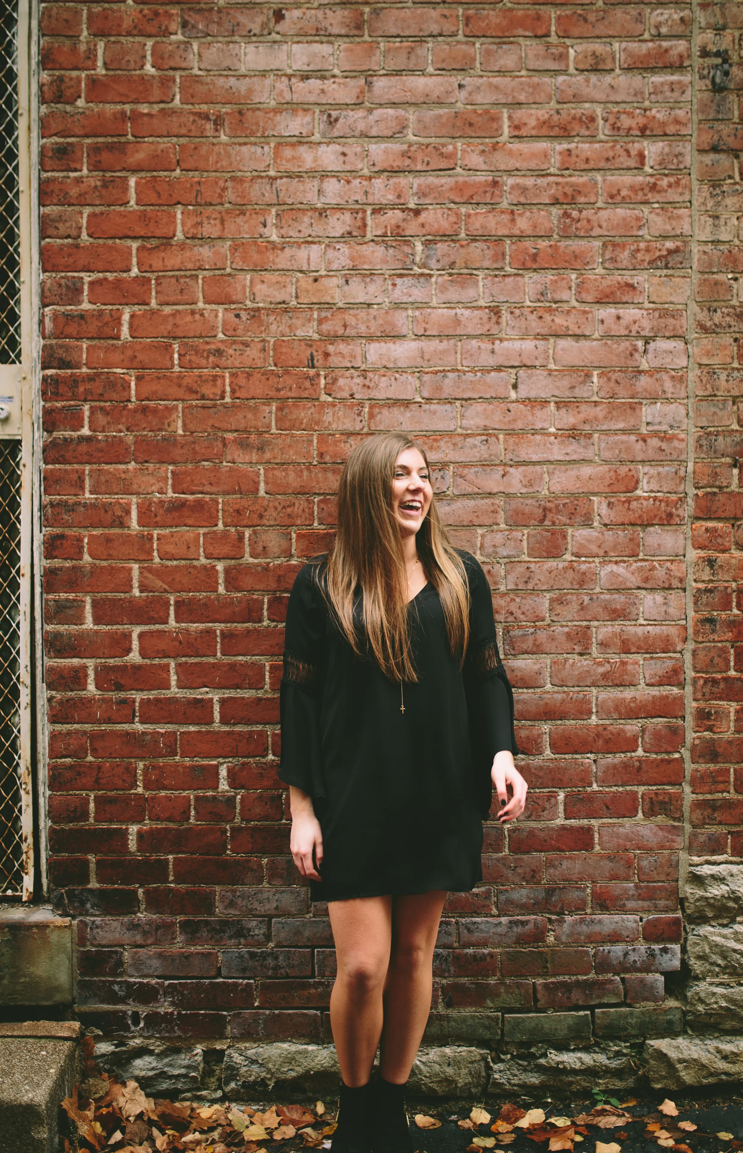 A young woman with long brown hair wearing a black dress stands in front of a red brick wall. She is smiling and looking to her left, with autumn leaves on the ground.