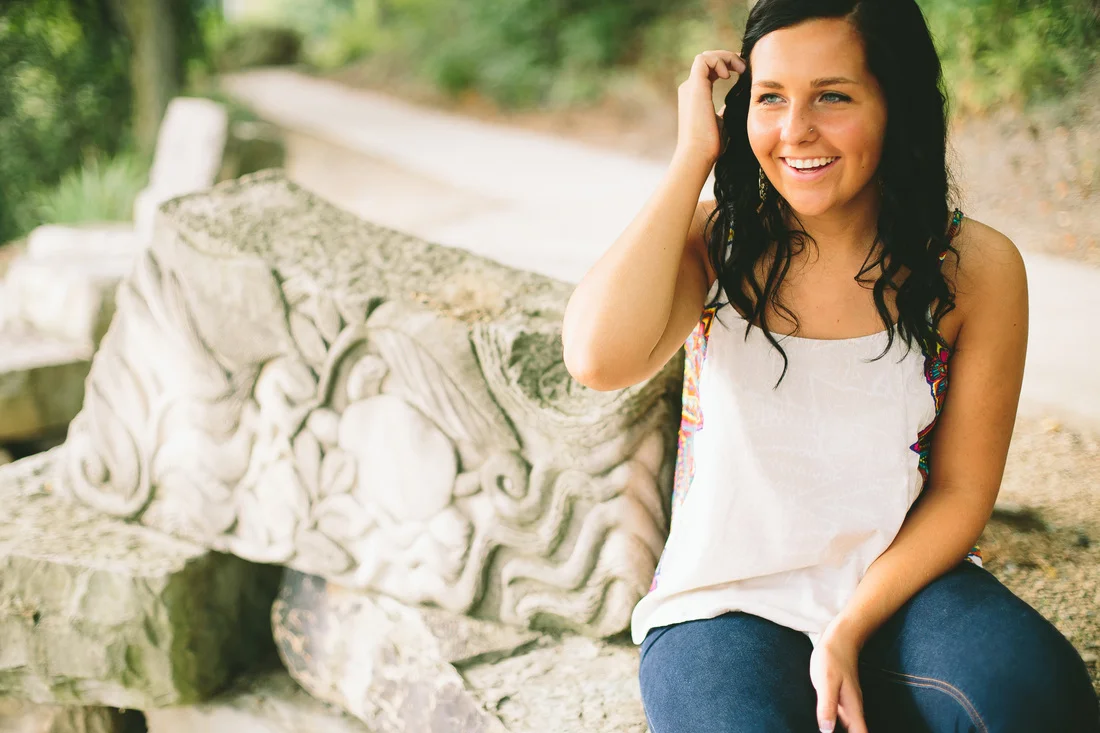 A woman with black hair and light skin sitting outdoors on a large, carved stone bench, smiling and touching her hair with her right hand, wearing a white tank top and dark jeans, with a greenery background.