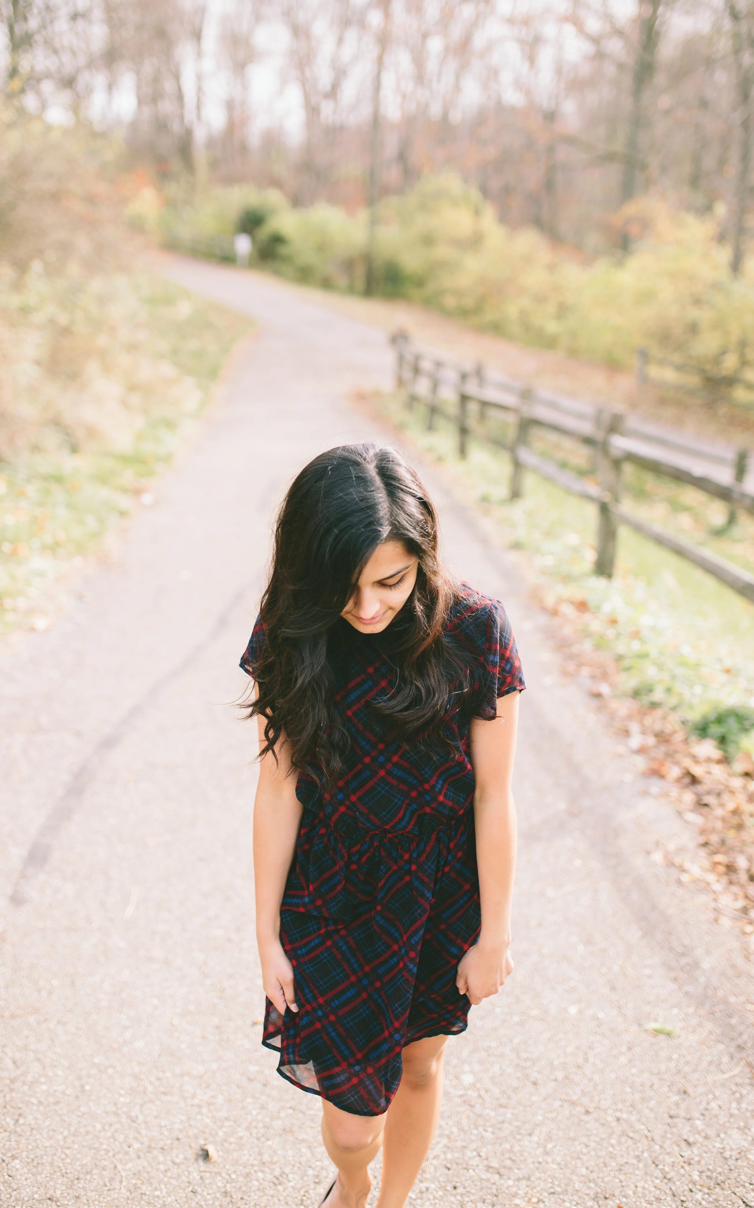 A young woman with long dark hair wearing a red plaid dress walking on a gravel path in a wooded park during autumn.