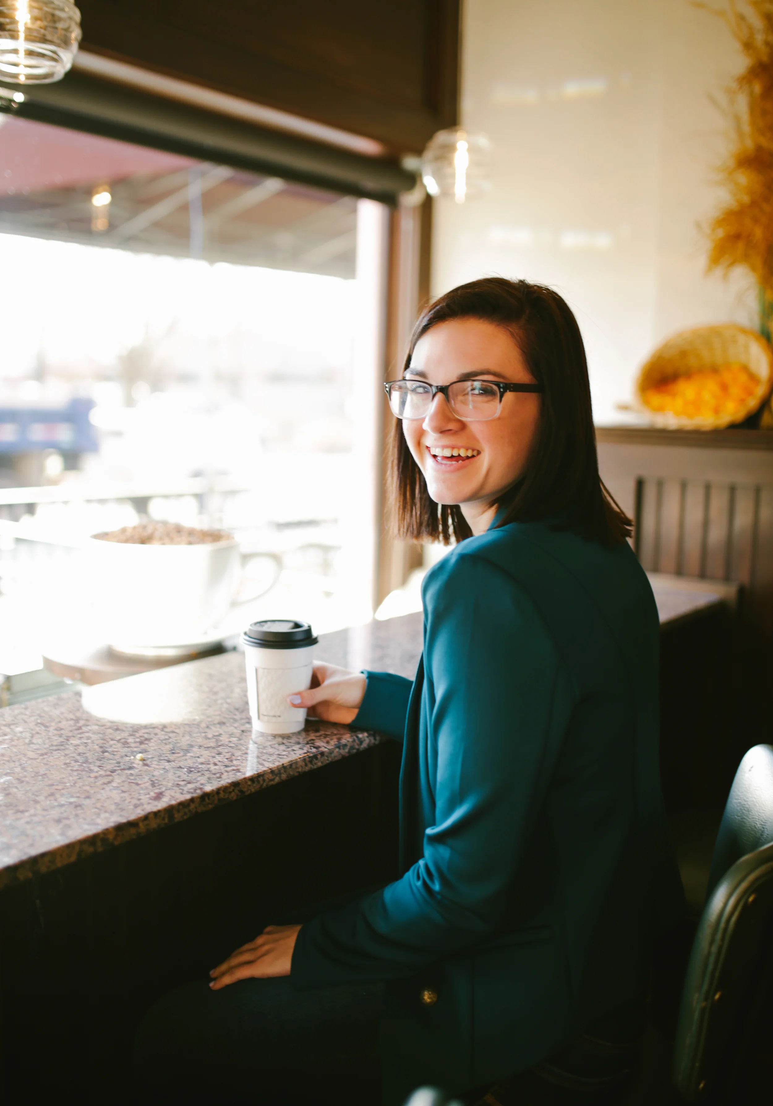 A woman with glasses and dark hair smiling while sitting at a counter in a coffee shop, holding a to-go coffee cup.