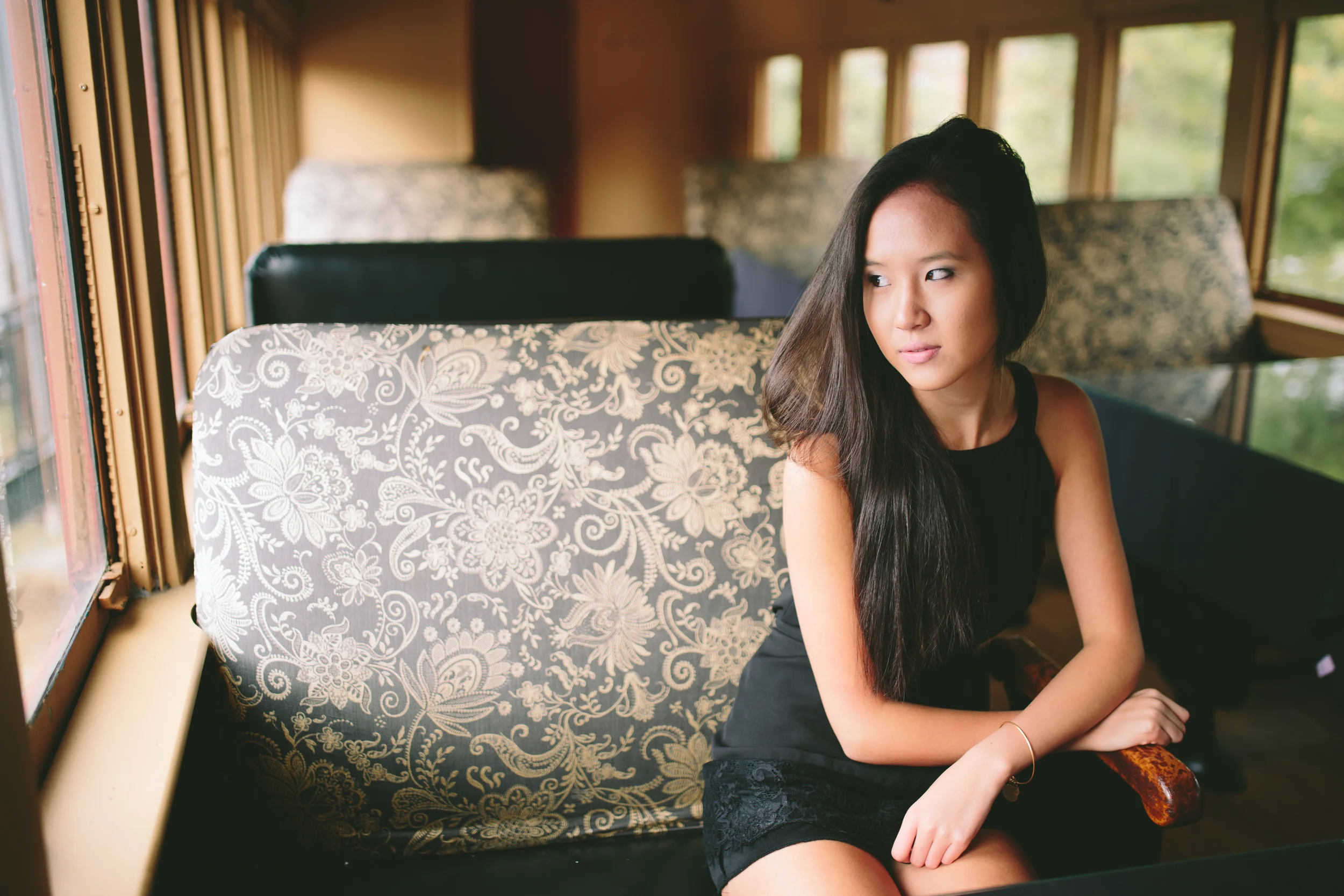 A young woman sitting on a vintage train with patterned seat covers, looking out the window with a contemplative expression.